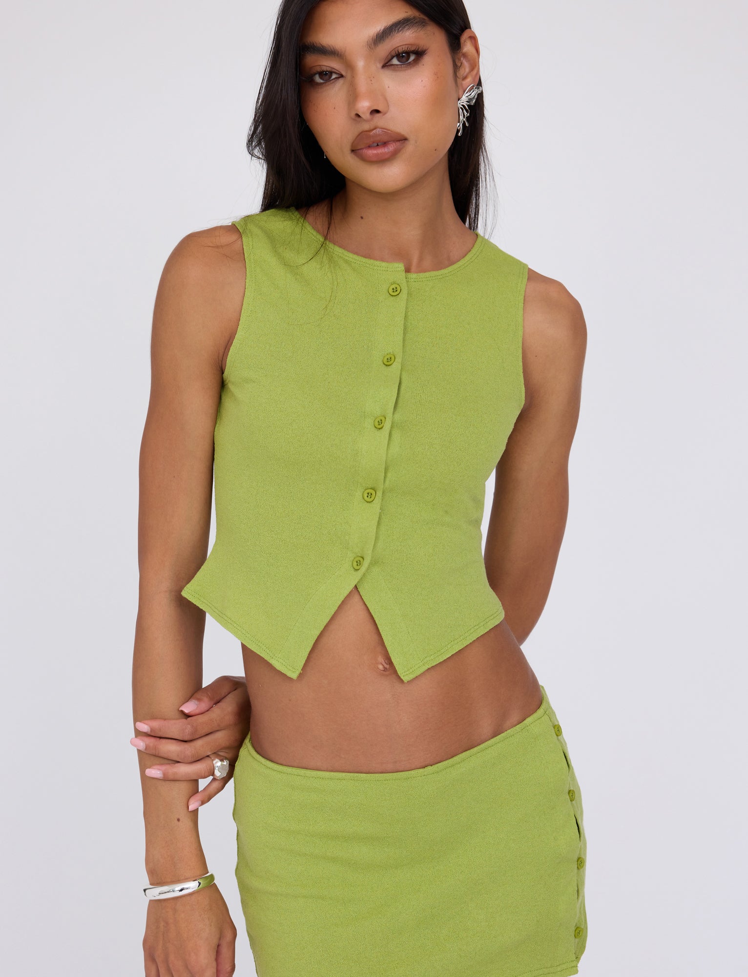 A person with long dark hair is wearing the OAKLEE TOP in Matcha Green from TIGERMIST, paired with a matching skirt featuring side buttons. They have light makeup with cat-eye eyeliner and are accessorized with earrings, bracelets, and rings. The background is plain white.