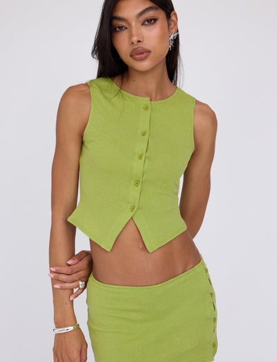 A person with long dark hair is wearing the OAKLEE TOP in Matcha Green from TIGERMIST, paired with a matching skirt featuring side buttons. They have light makeup with cat-eye eyeliner and are accessorized with earrings, bracelets, and rings. The background is plain white.