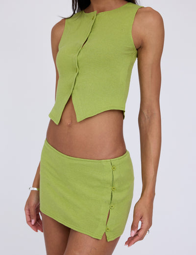 A person wearing the OAKLEE TOP - GREEN : MATCHA GREEN from TIGERMIST, a green sleeveless crop top with a front opening, paired with a matching green mini skirt featuring button details on one side. The outfit reveals the midriff and is complemented by simple wrist jewelry. The background is plain white.