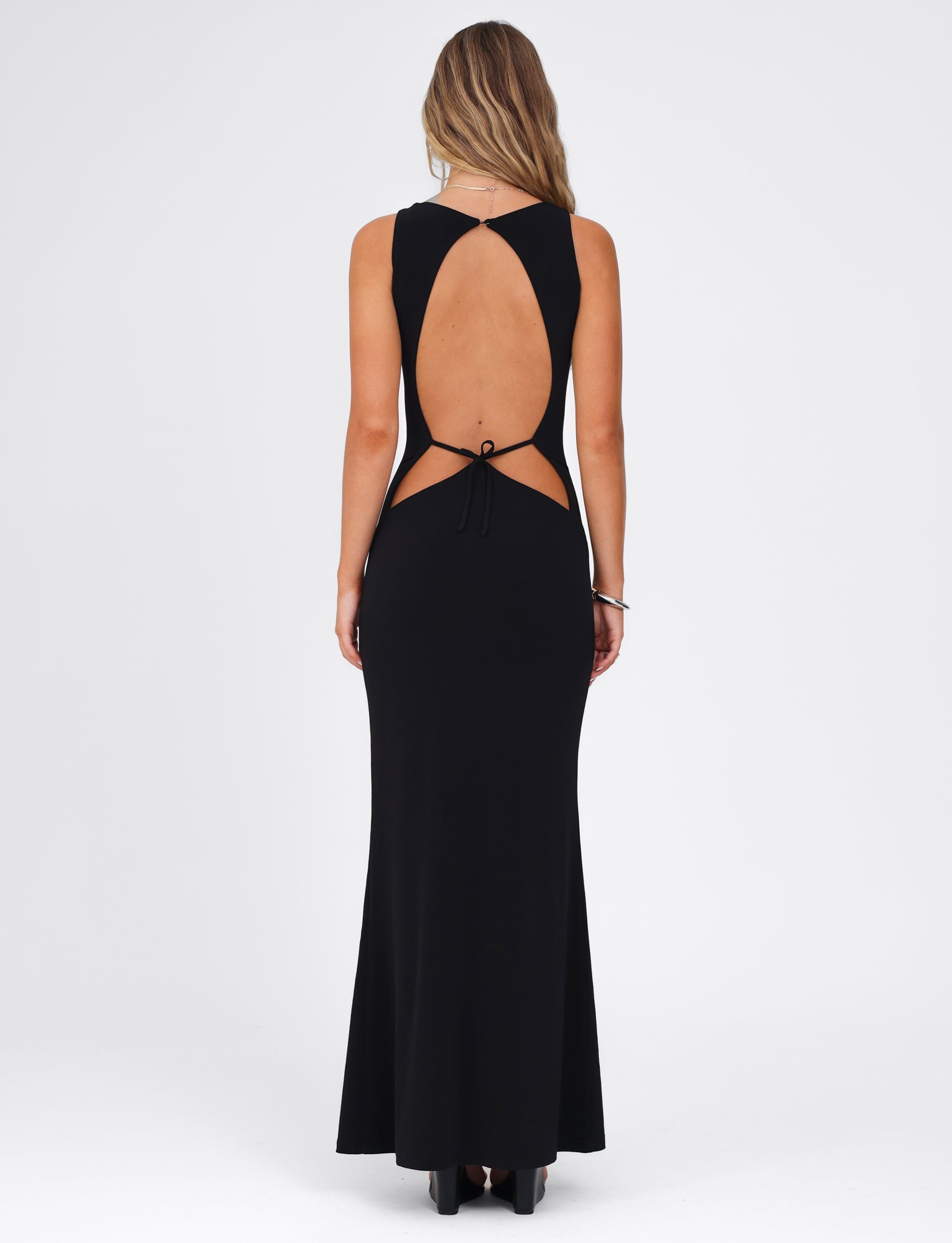 A woman, with her back to the camera, is elegantly dressed in the Sonnet Maxi Dress - Black by Tigermist. This sophisticated long black gown showcases a chic open-back design accompanied by a tie detail. Her long, wavy hair flows down as she gracefully stands in black high-heeled shoes.