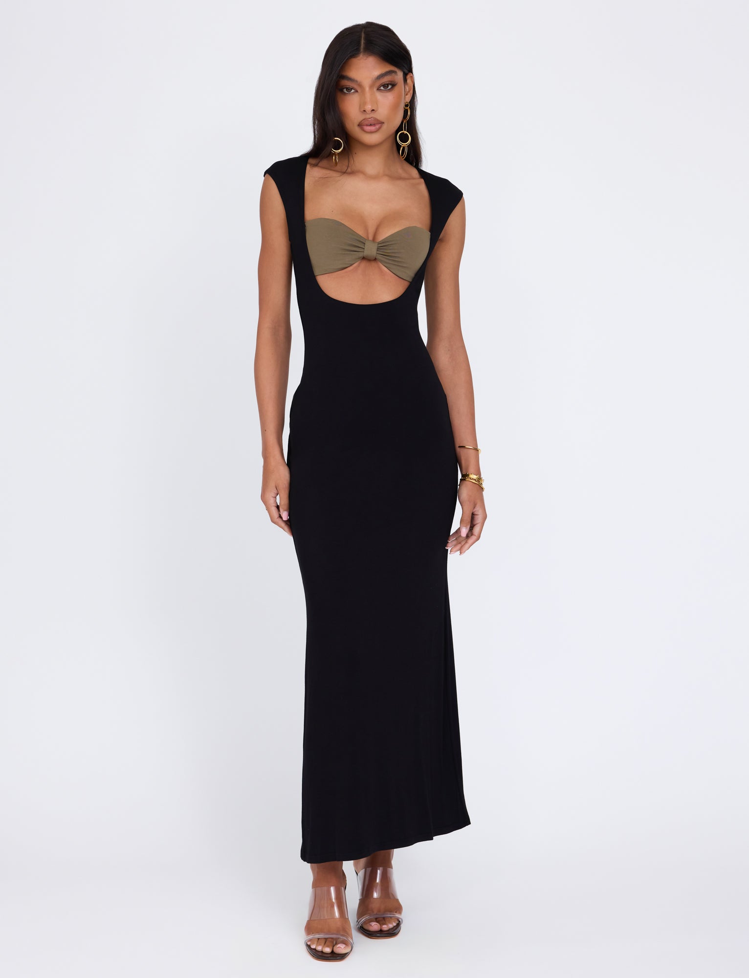 A woman stands confidently in the TIGERMIST BLAKELY DRESS - BLACK, a form-fitting, floor-length dress featuring a cut-out bodice with an olive green bandeau top underneath. She accessorizes with hoop earrings, a bracelet, and strappy heels against a plain white background.