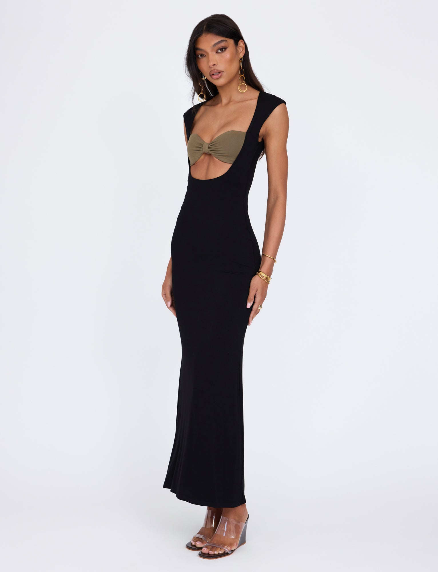 A woman with long hair is wearing the Blakely Dress in black by TigerMist, featuring a plunging neckline that reveals a tan bandeau underneath. She accessorizes with gold hoop earrings, bracelets, and clear high-heeled sandals while standing against a plain white background.