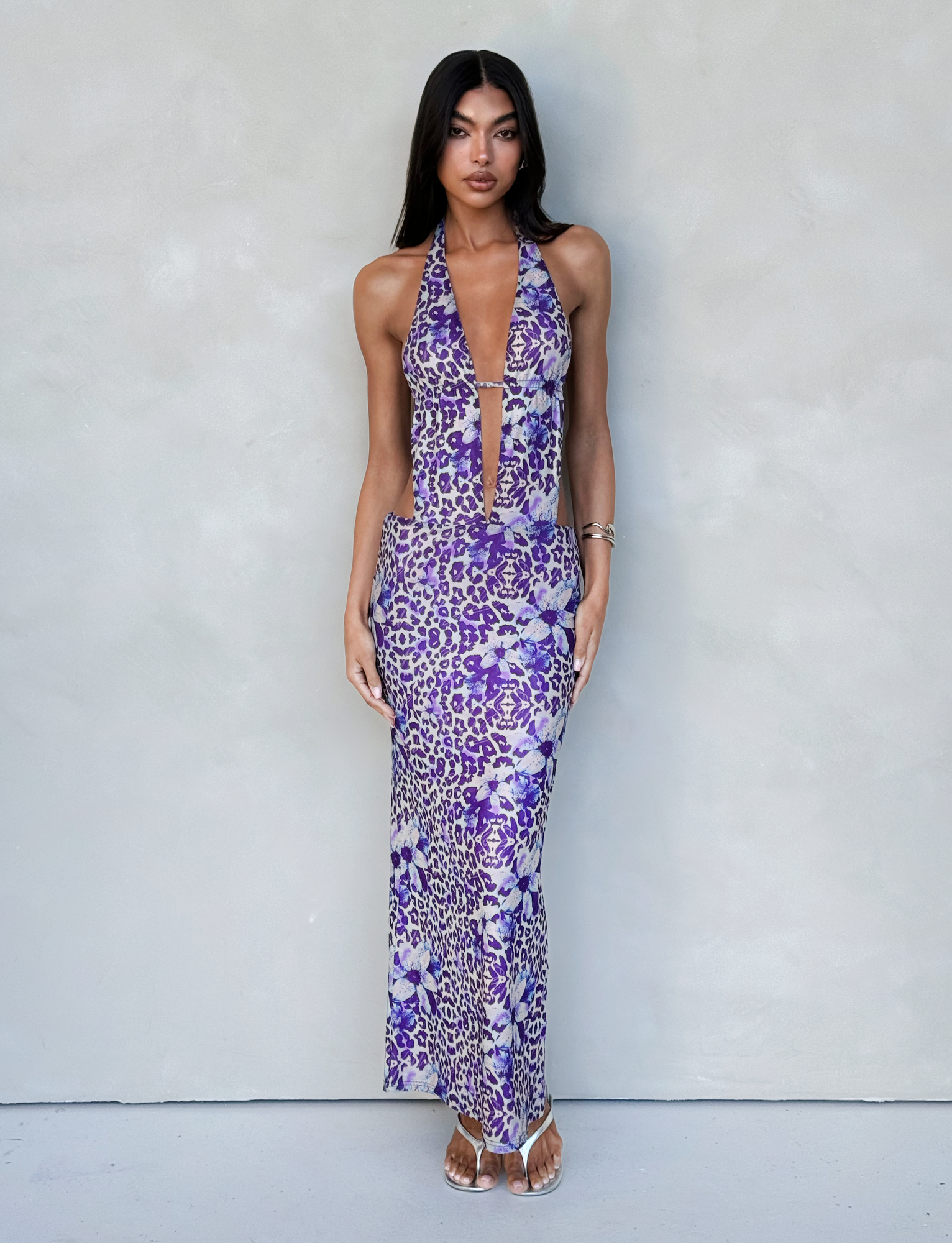 A woman with long dark hair stands against a neutral gray wall, wearing the TIGERMIST MAGDALENA MAXI DRESS - PURPLE : BY POPPY LEOPARD, a floor-length halter-neck dress featuring a purple animal and floral print. Her attire boasts a deep V-neckline. She accessorizes with bracelets and wears strappy, transparent high heels.