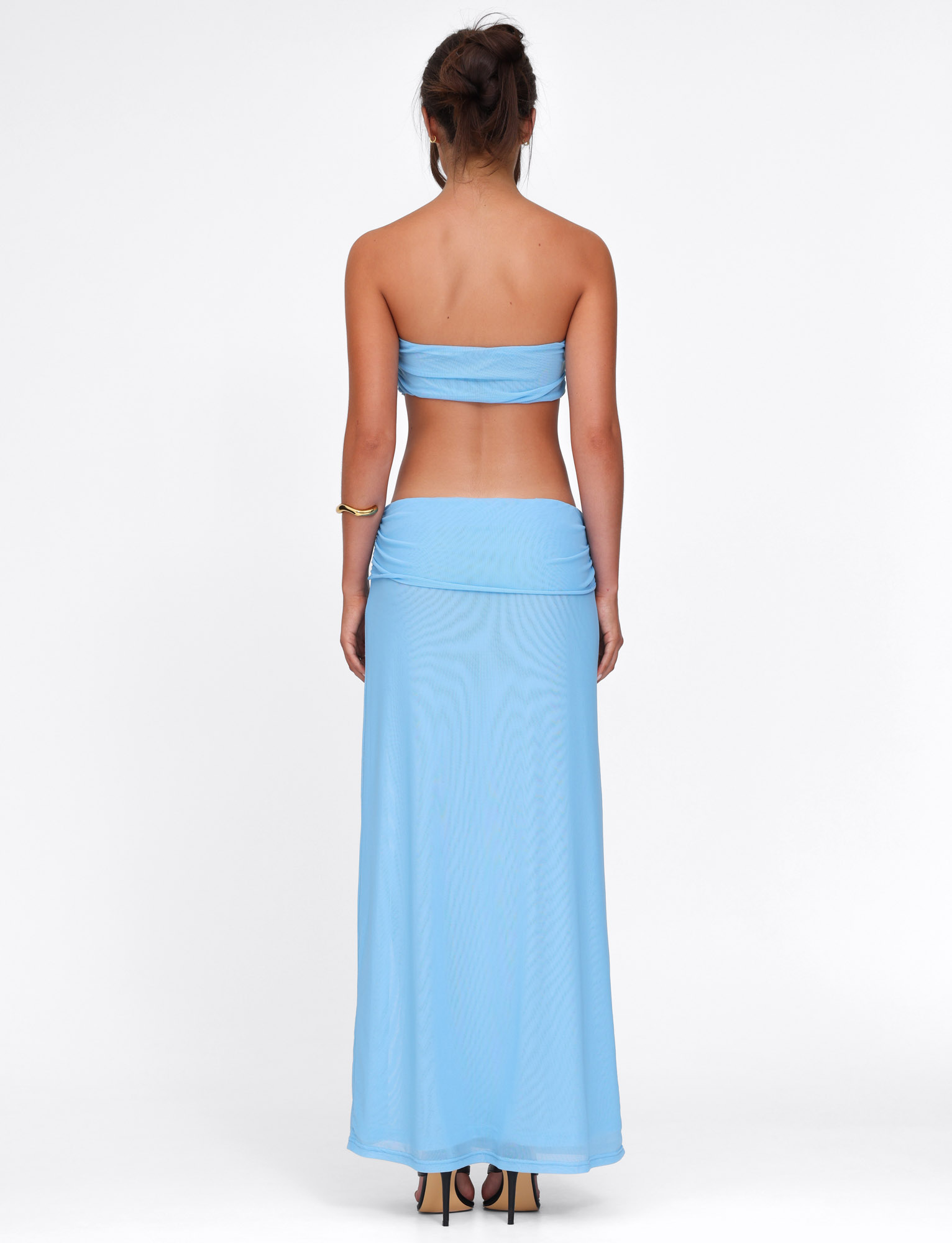 A woman with her hair up stands facing away, wearing the TIGERMIST KYNLEE DRESS in sky blue—a strapless, mesh-fabricated long dress—paired with a gold bracelet, set against a plain white background.
