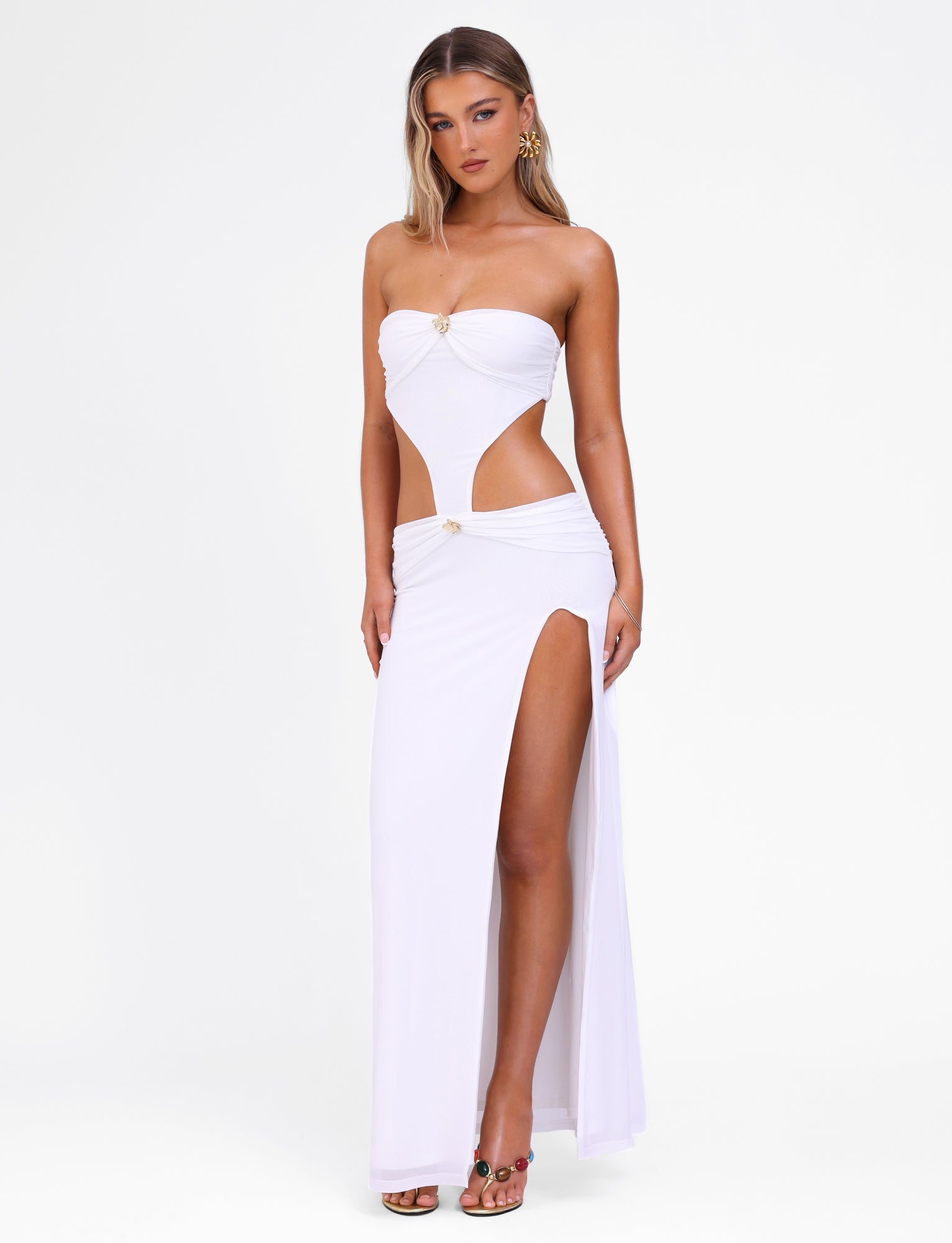 A woman is wearing the TIGERMIST KYNLEE MAXI DRESS - WHITE, a strapless evening gown featuring cutout details at the sides and a high slit on one leg. She stands against a plain white background, accessorized with floral gold metal hardware hoop earrings and open-toe heels. Her hair is styled in loose waves.