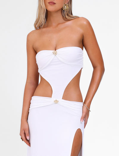 A woman wearing the TIGERMIST KYNLEE MAXI DRESS - WHITE, an elegant, strapless white ensemble with mesh fabrication and a unique cut-out design at the midriff, coupled with a high slit. She accessorizes with floral gold metal earrings and bracelets, complemented by her blonde hair cascading over her shoulders.