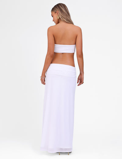A woman with long blonde hair is standing and facing away from the camera, wearing a strapless KYNLEE MAXI DRESS - WHITE by TIGERMIST, along with a matching long skirt. Adorned with floral gold metal hardware, she has a bracelet on her wrist and stands against a plain white background.