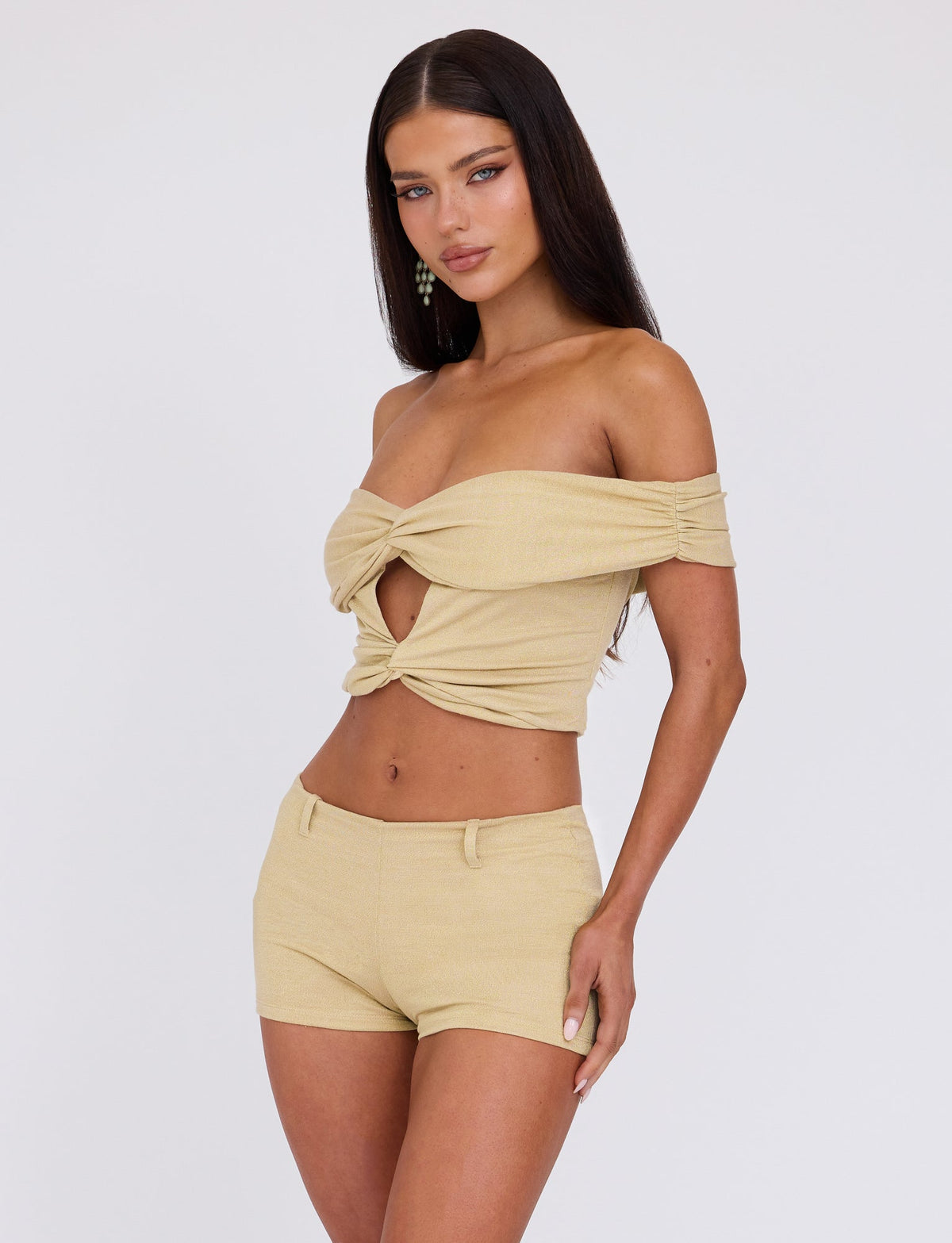A woman with long dark hair is posing against a plain white background. She is wearing the LYNSEY TOP - GOLD from TIGERMIST, a light beige off-shoulder crop top with a twist knot in the front, paired with matching high-waisted shorts. Her right hand is resting on her hip, and she is looking directly at the camera.