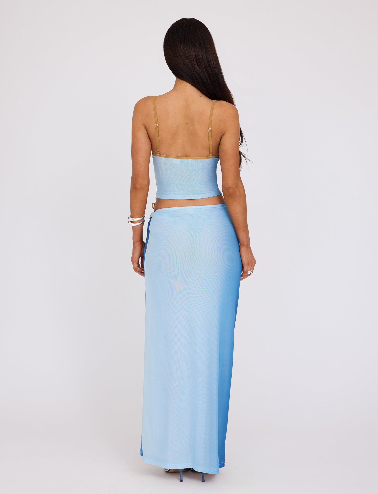A woman with long, dark hair stands facing away from the camera, showcasing a TigerMist ELVERA SKIRT - BLUE : GRADIENT FLORAL two-piece outfit with a crop top and a high-waisted, ankle-length skirt. The top has thin shoulder straps, and she accessorizes with white bracelets on her left wrist.