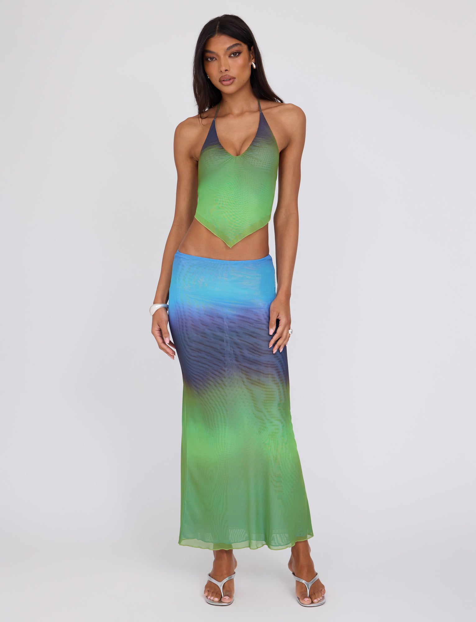 A woman with long dark hair is modeling the TIGERMIST NOELY TOP - MULTI : OMBRE : GRADIENT STRIPE halter-neck top and a long skirt featuring a gradient design that transitions from green to blue and purple. She is standing against a plain background, wearing strappy white sandals and minimal jewelry.