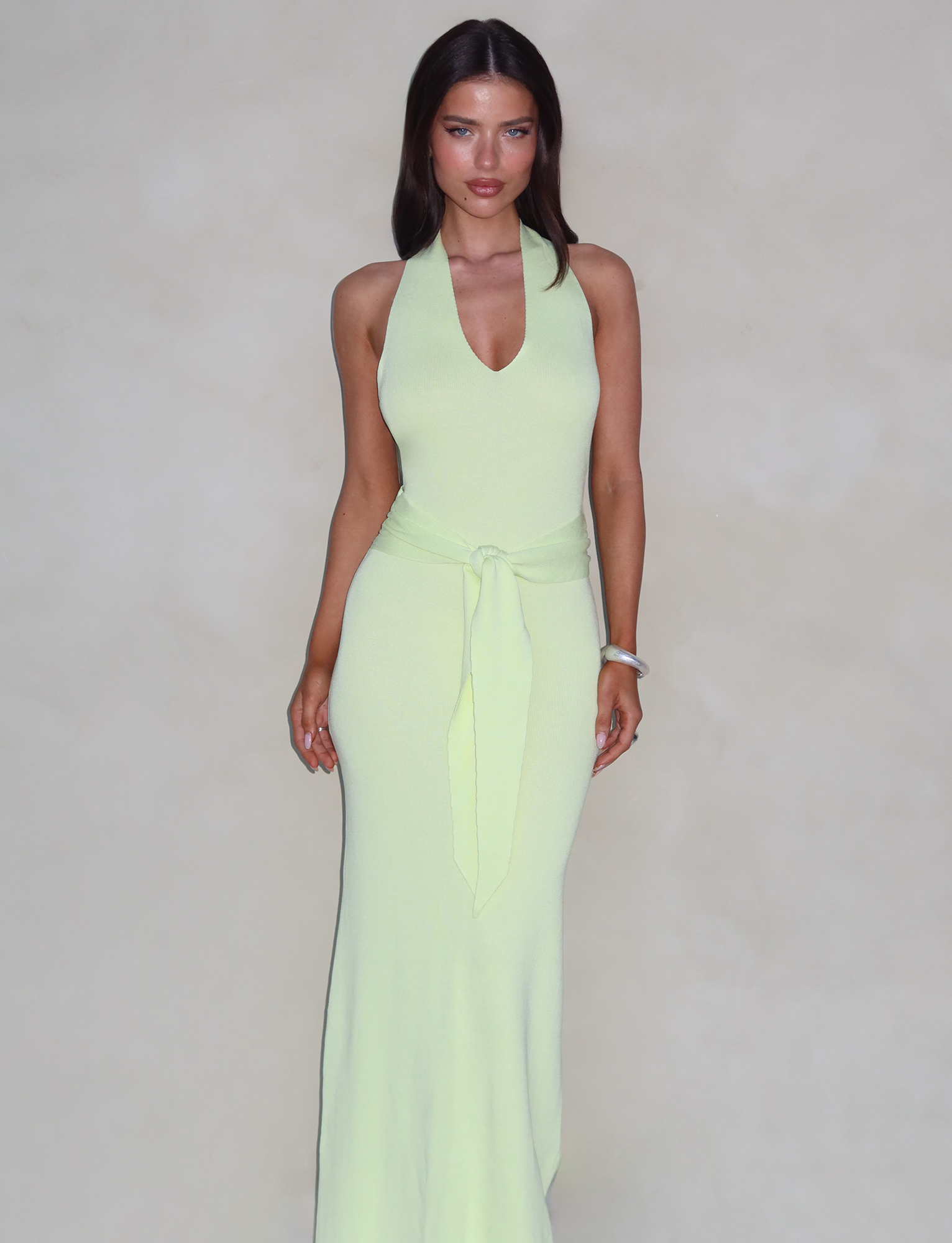 A woman with straight, dark hair is wearing the TIGERMIST MAREE DRESS in green, which features a V-neck and a tie at the waist. The sleeveless, maxi-length dress highlights her elegant silhouette as she stands against a neutral, beige background, looking directly at the camera with a neutral expression.