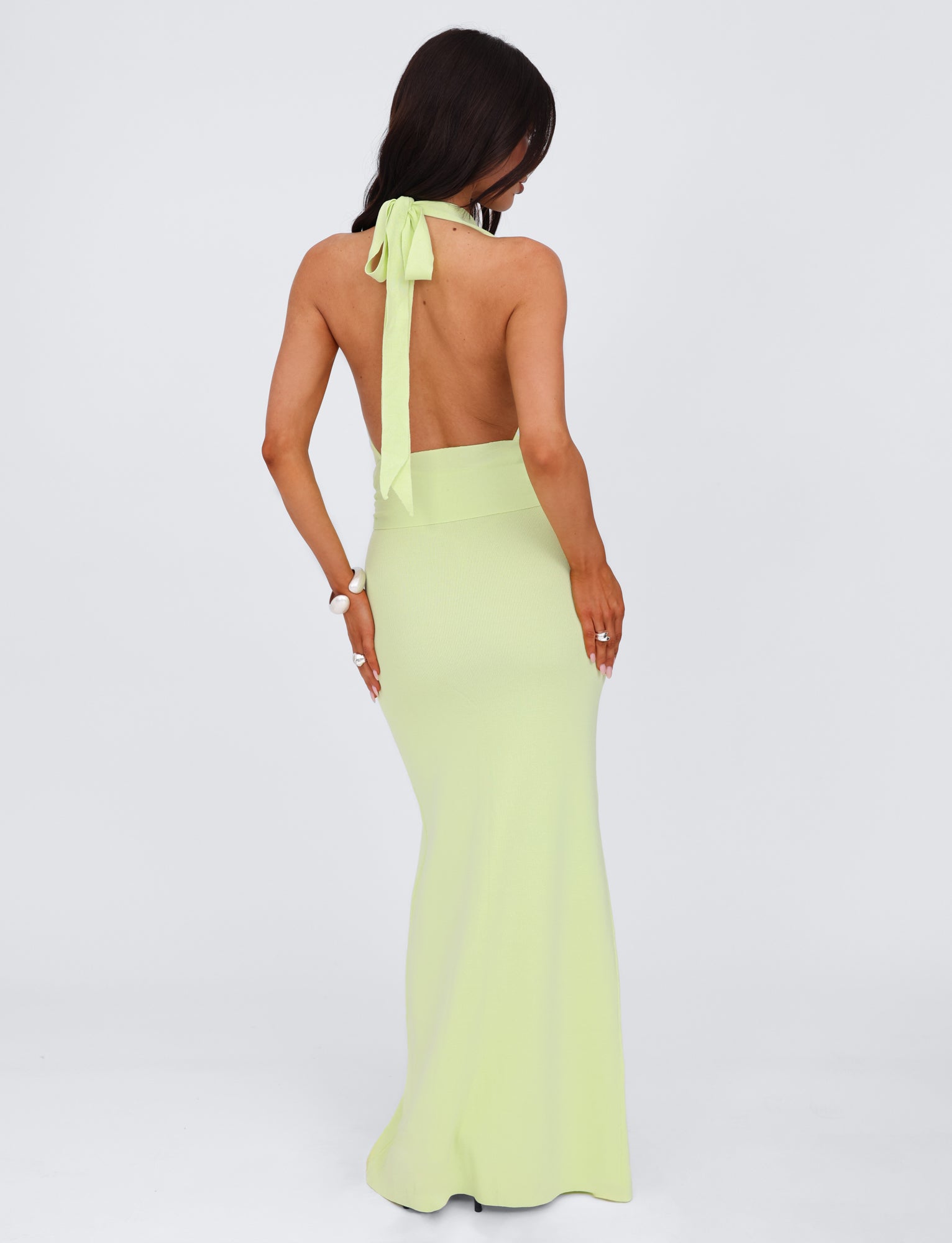 A woman with long brown hair is wearing the MAREE DRESS - GREEN by TIGERMIST, a backless light green knit halter dress. She faces away from the camera, showcasing the open back and maxi length of the dress as she stands against a plain white background.