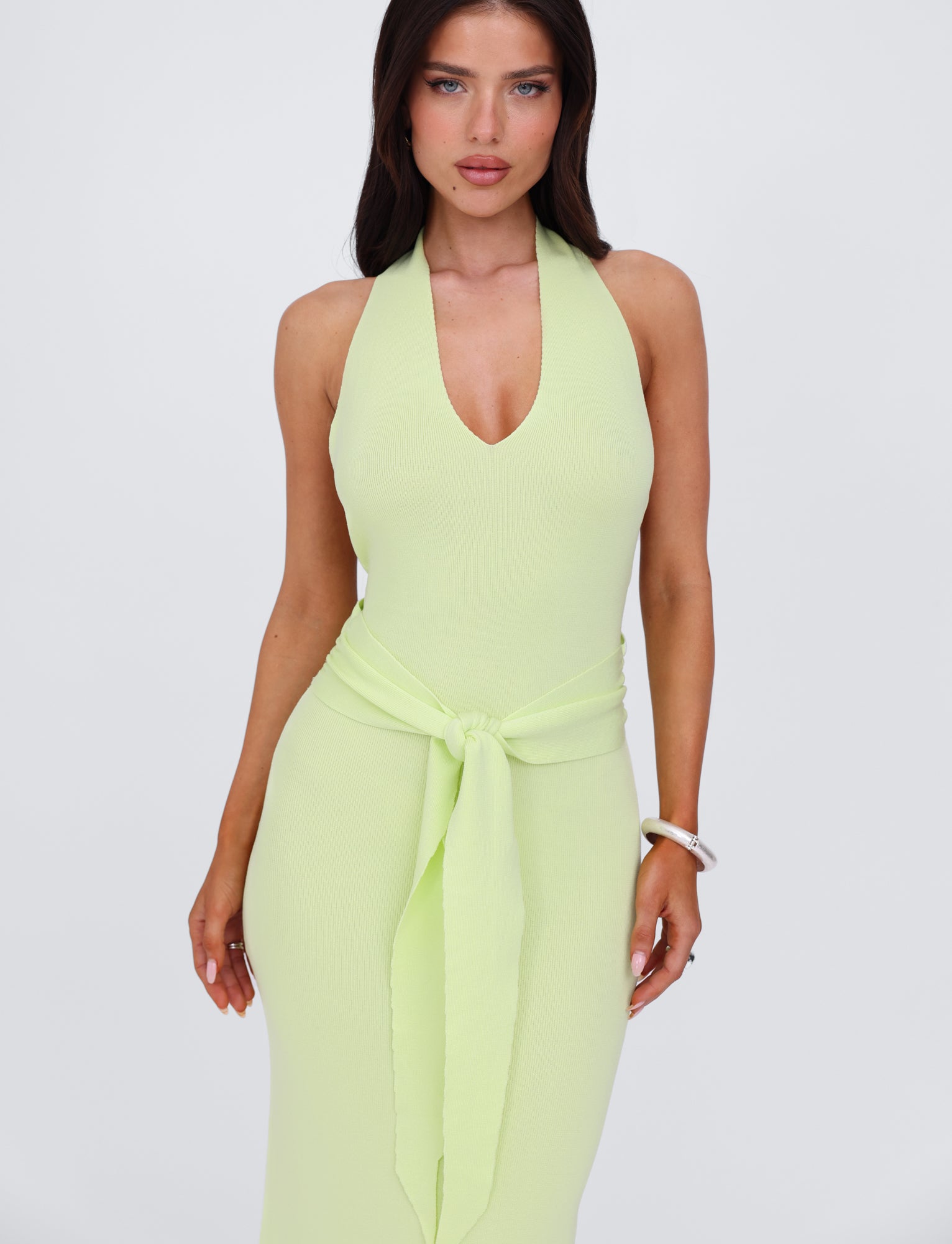 A woman with long brown hair is wearing the MAREE DRESS - GREEN by TIGERMIST, a light green knit dress in a halter style with a knotted belt at the waist. She stands against a plain white background, one arm resting by her side and the other slightly bent. A silver bracelet adorns her right wrist.