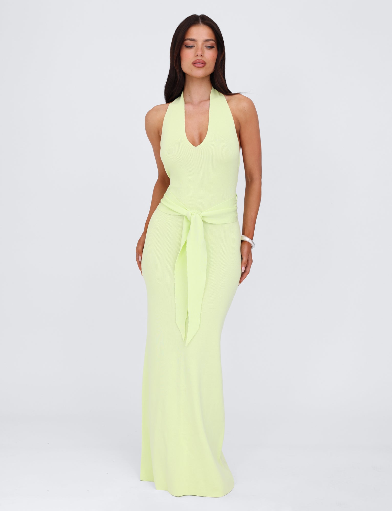 A woman stands against a plain background wearing the MAREE DRESS - GREEN by TIGERMIST, a sleeveless light yellow-green knit halter neck dress with a deep V-neckline and tied fabric detail at the waist. She has long, dark hair and accessorizes with minimal jewelry.