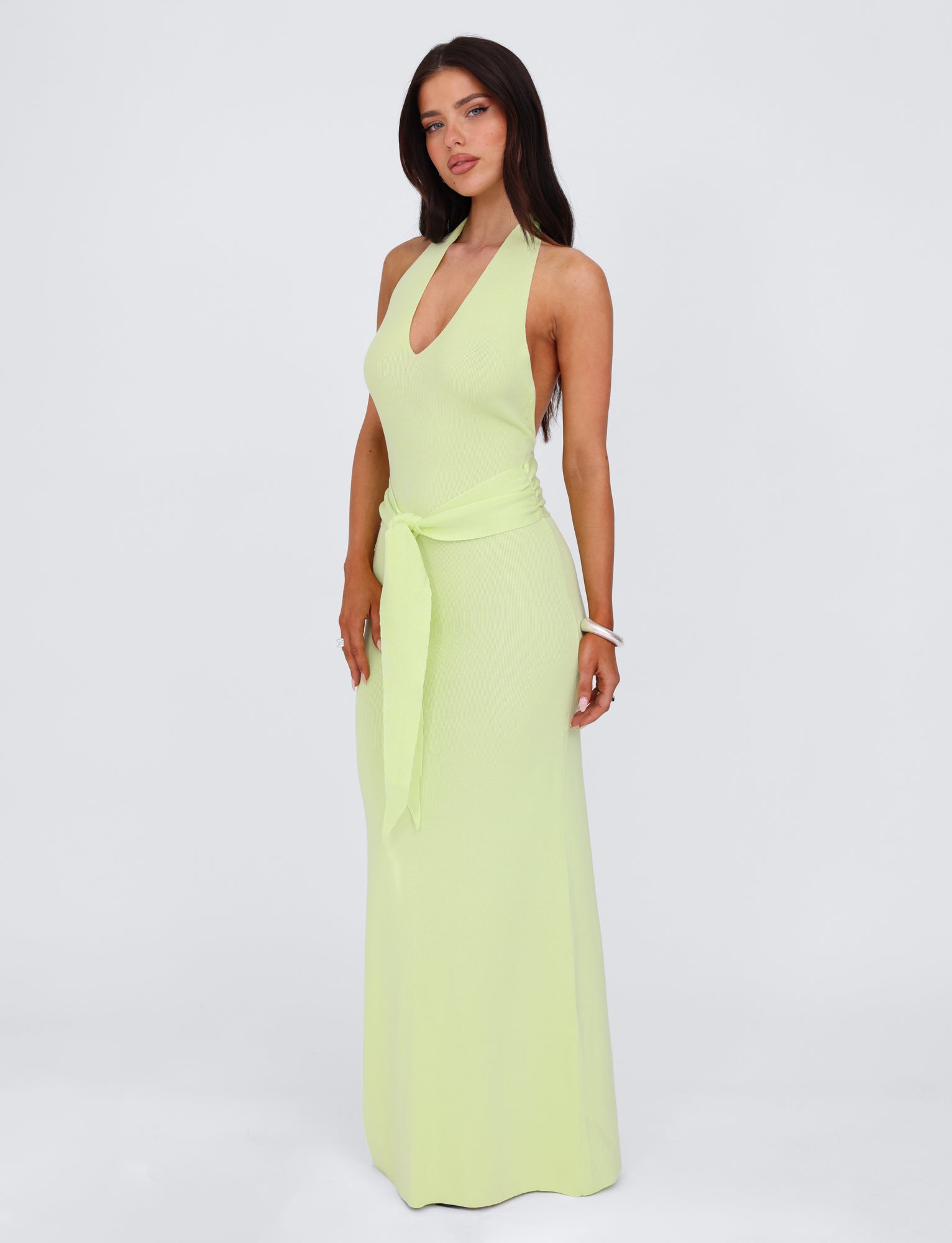 A woman stands against a plain white background, wearing the TIGERMIST MAREE DRESS in green. The long knit dress features a deep V-neck and a tied belt at the waist, coming down to maxi length. Her long, dark hair is highlighted by the sleeveless design. She accessorizes with a white bracelet on her right wrist and gazes straight ahead.
