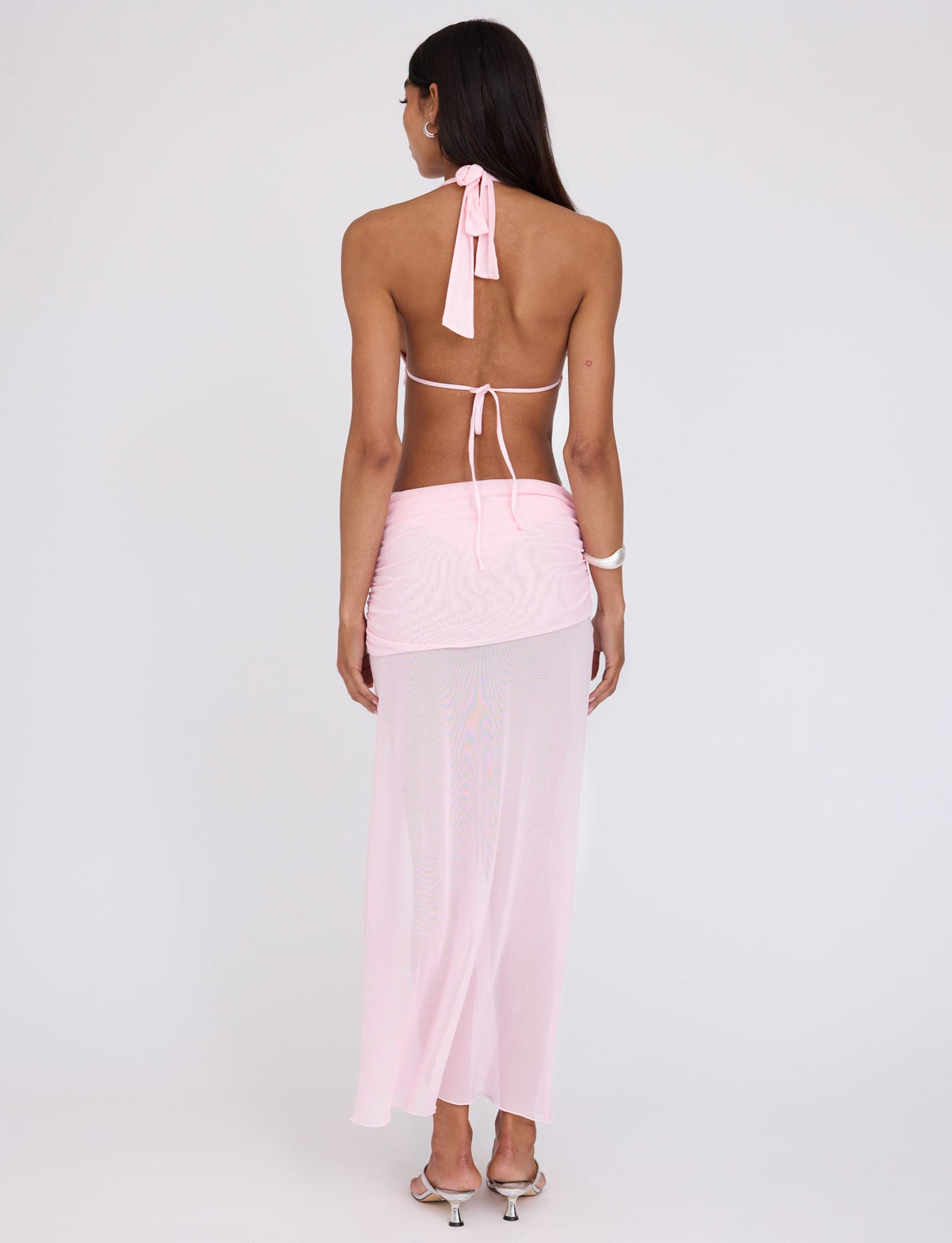 A person with long dark hair stands facing away, wearing a light pink ESHA MAXI DRESS from TIGERMIST. They also have on silver high-heeled sandals and a white bracelet on their left wrist. The background is plain and neutral.