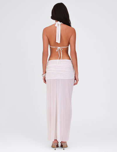 Rear view of a person with long dark hair, wearing the TIGERMIST ESHA MAXI DRESS - WHITE, featuring a halter-style bikini top paired with a sheer, full-length skirt. They are standing against a plain, light background.