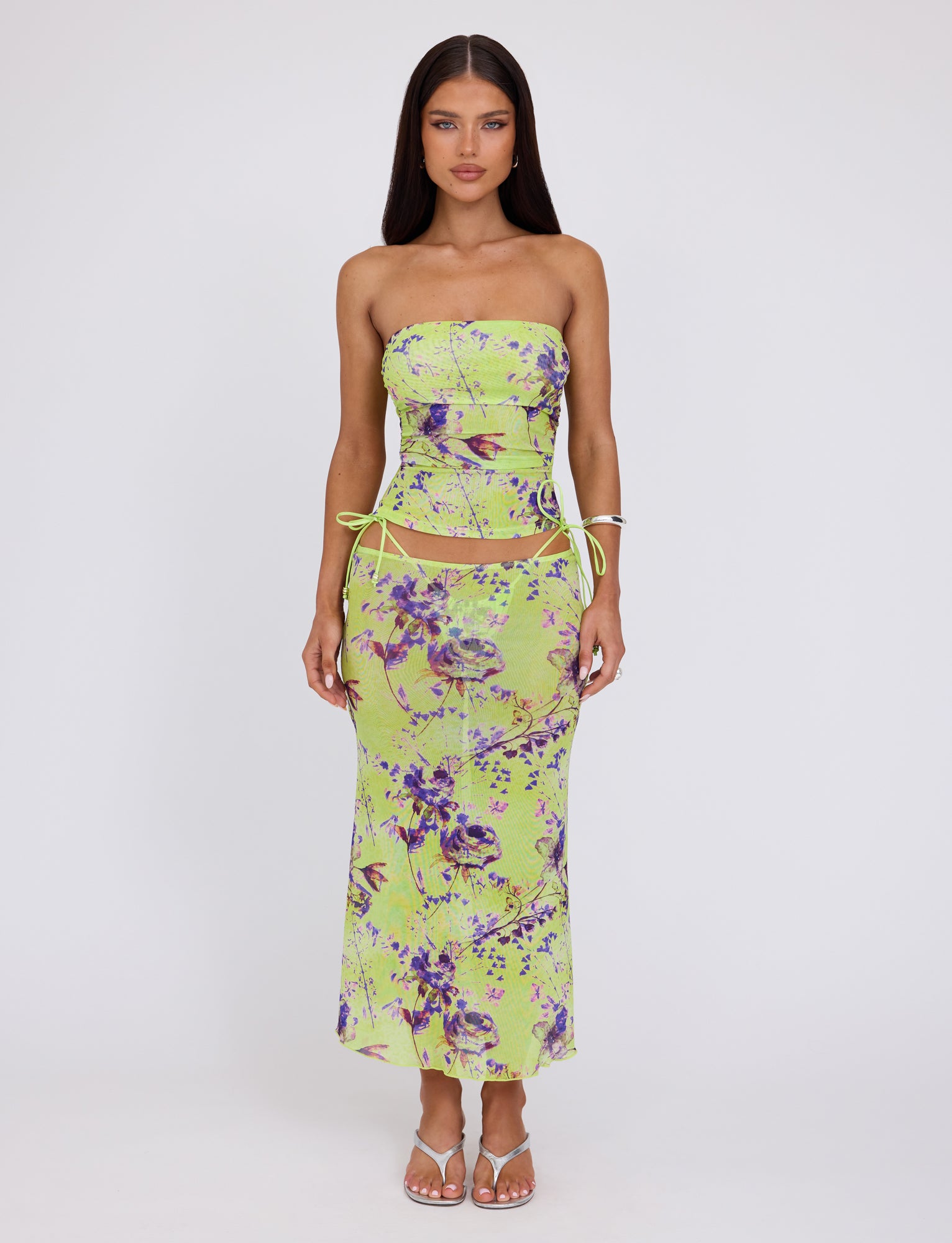 A woman with long dark hair stands against a plain white background. She is wearing the Kellan Top in green by Tigermist, featuring a beautiful Poppy Floral design. The top has stylish side-ties with cutouts around the waist, and she complements her look with white sandals.