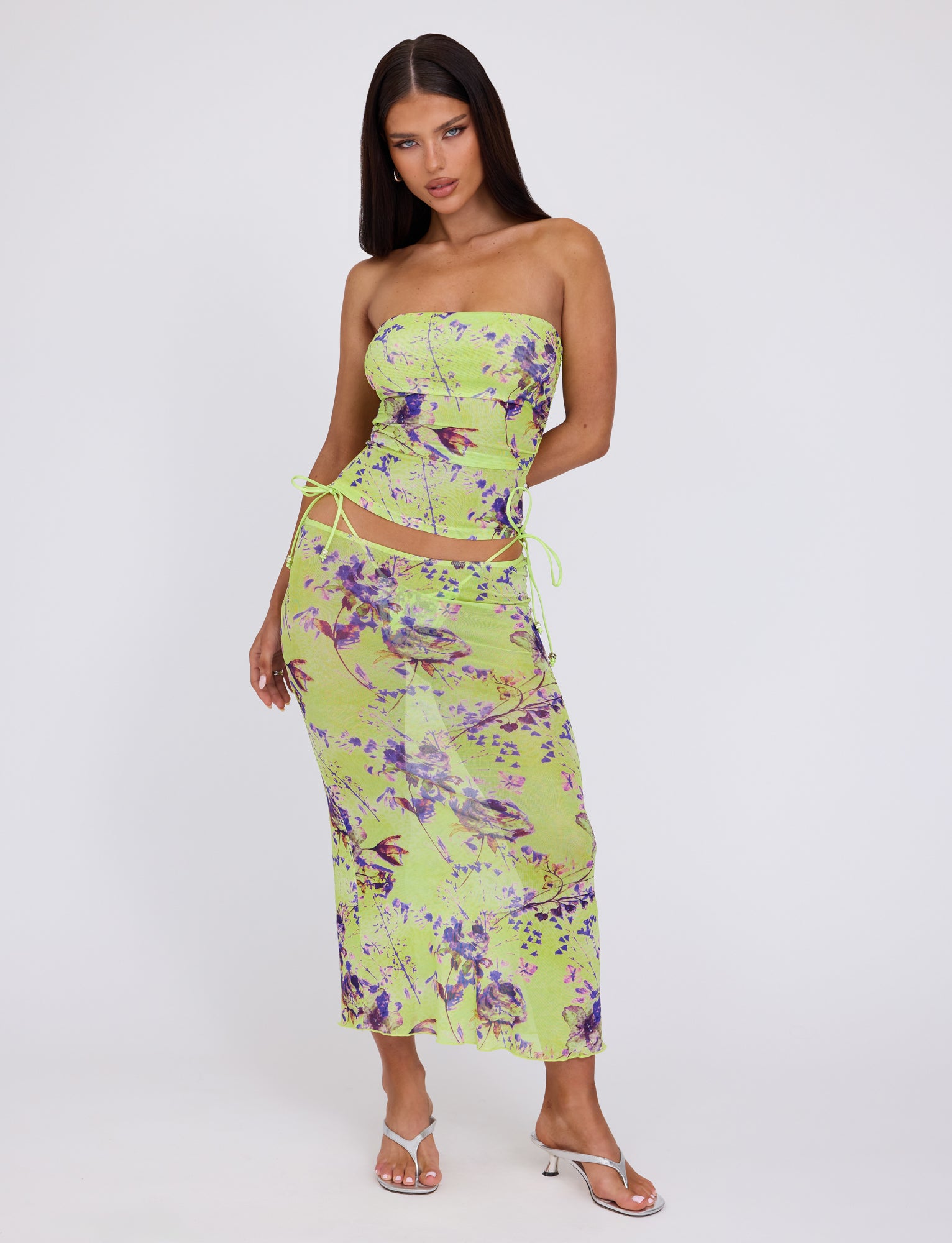 A woman with long dark hair models the Kellan Top - Green by Poppy Floral from Tigermist, featuring a vibrant green and purple abstract pattern. The top is tied at the sides with thin straps, and she pairs it with white high-heeled sandals. She stands against a plain white background.