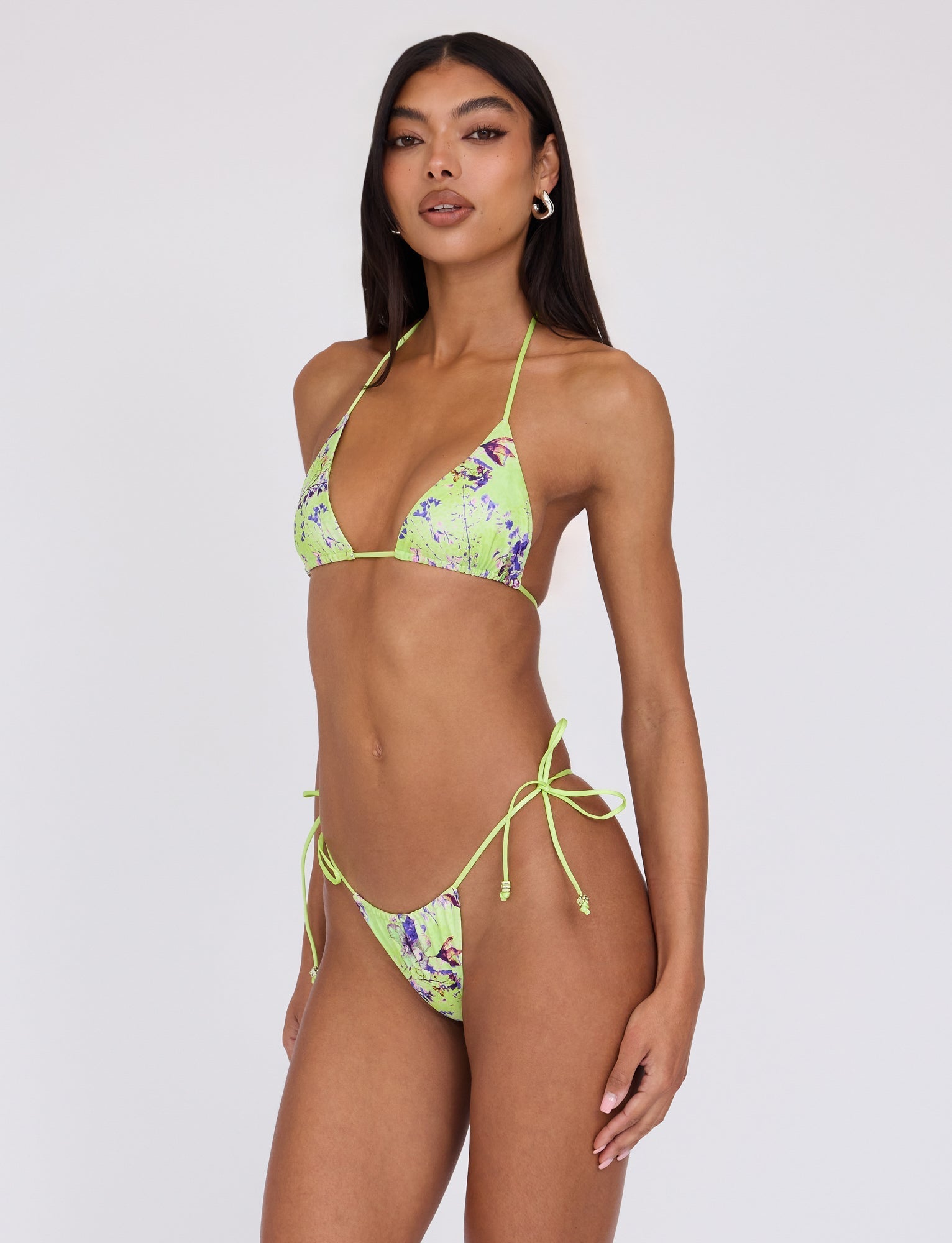 A person with long, dark hair is wearing the AVE Bikini Top - Green by Poppy Floral from TIGERMIST. The bikini has a vibrant floral design and is tied with thin straps around the neck and back, while the matching bottom is tied at the sides. The individual stands against a plain, light background.