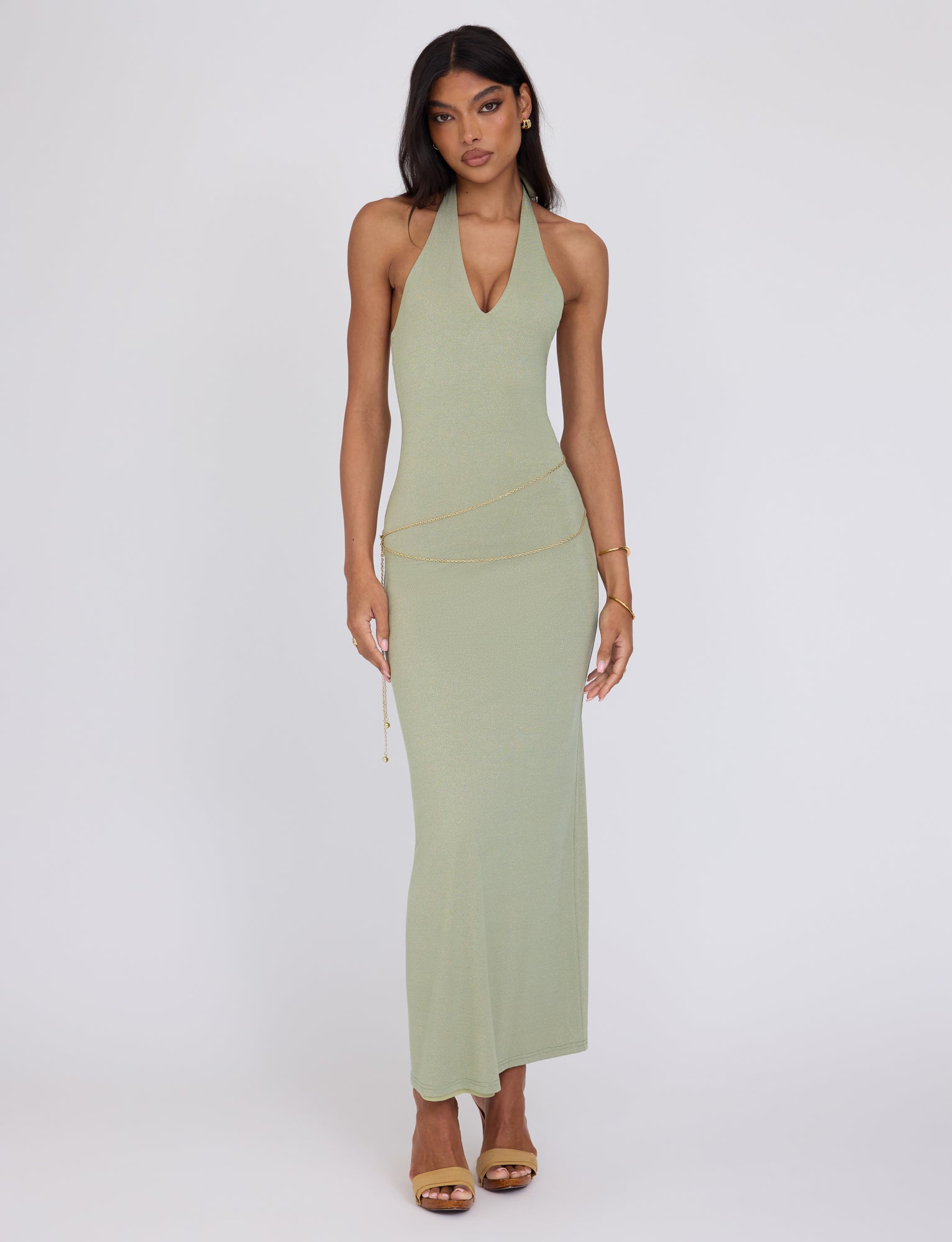 A woman stands against a plain background wearing a light green TIGERMIST Opeli Maxi Dress in Green Sparkle. She has long, straight hair and is accessorized with gold earrings, a delicate gold chain belt at the waist, and gold high-heeled sandals. Her expression is neutral, and her arms rest by her sides.