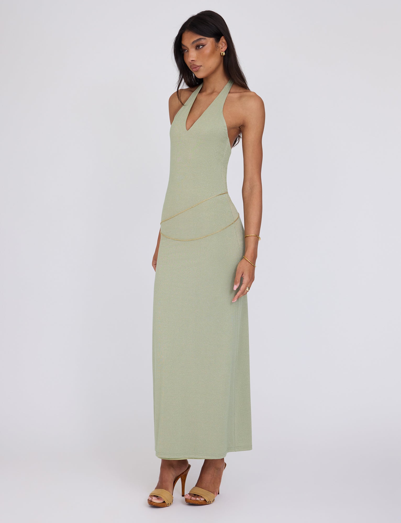 A woman wearing the Tigermist Opeli Maxi Dress in Green Sparkle, featuring a halter-neck and backless design, with a subtle gold chain belt, matching gold bracelet, and sandals stands against a plain white background. She has long, dark hair and a neutral expression.