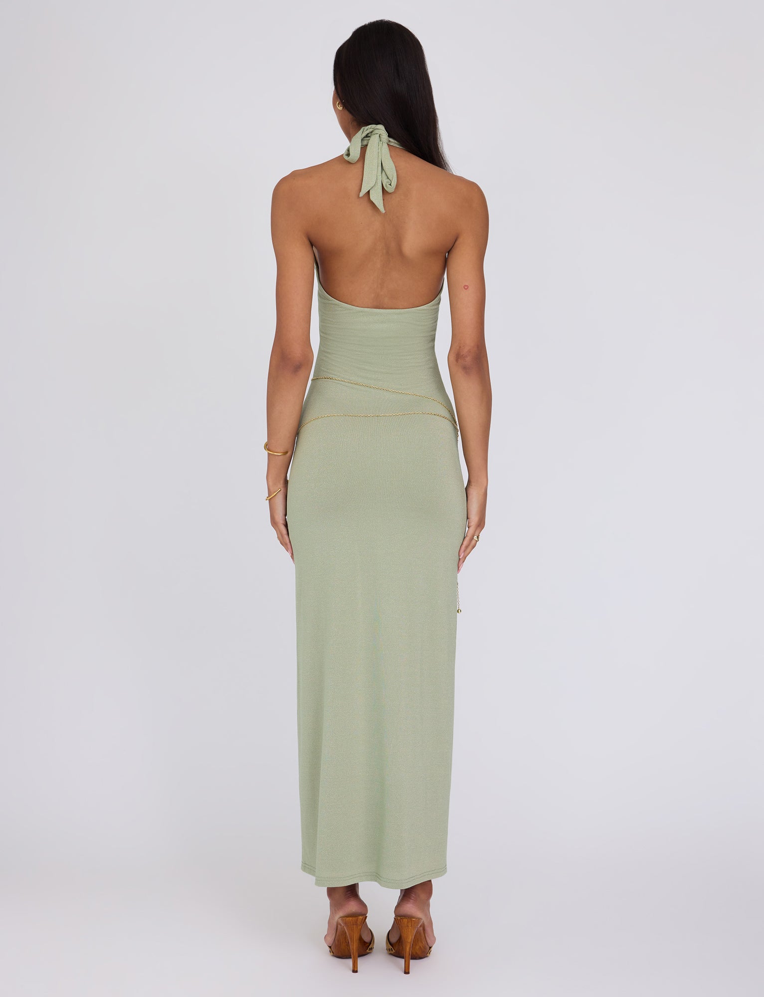 A person with long, dark hair is standing with their back to the camera, wearing a light green Opeli Maxi Dress - Green Sparkle from Tigermist that ties behind the neck. They appear to be wearing high-heeled sandals and a gold chain belt around their waist. A bracelet adorns their left wrist against the plain white background.