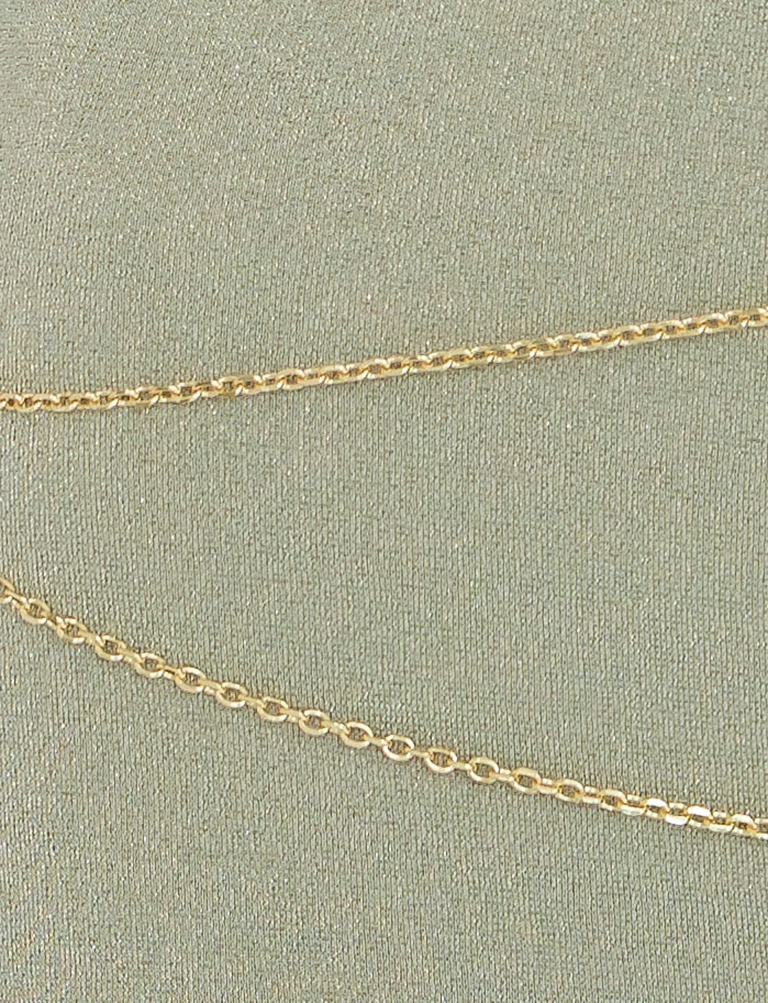 Close-up of a delicate gold chain necklace laid on the textured, light green fabric background of the TIGERMIST OPELI MAXI DRESS in Green Sparkle. The chain features small, linked oval-shaped links that reflect light subtly, adding a touch of elegance reminiscent of an opulent gold belt adorning this exquisite dress.