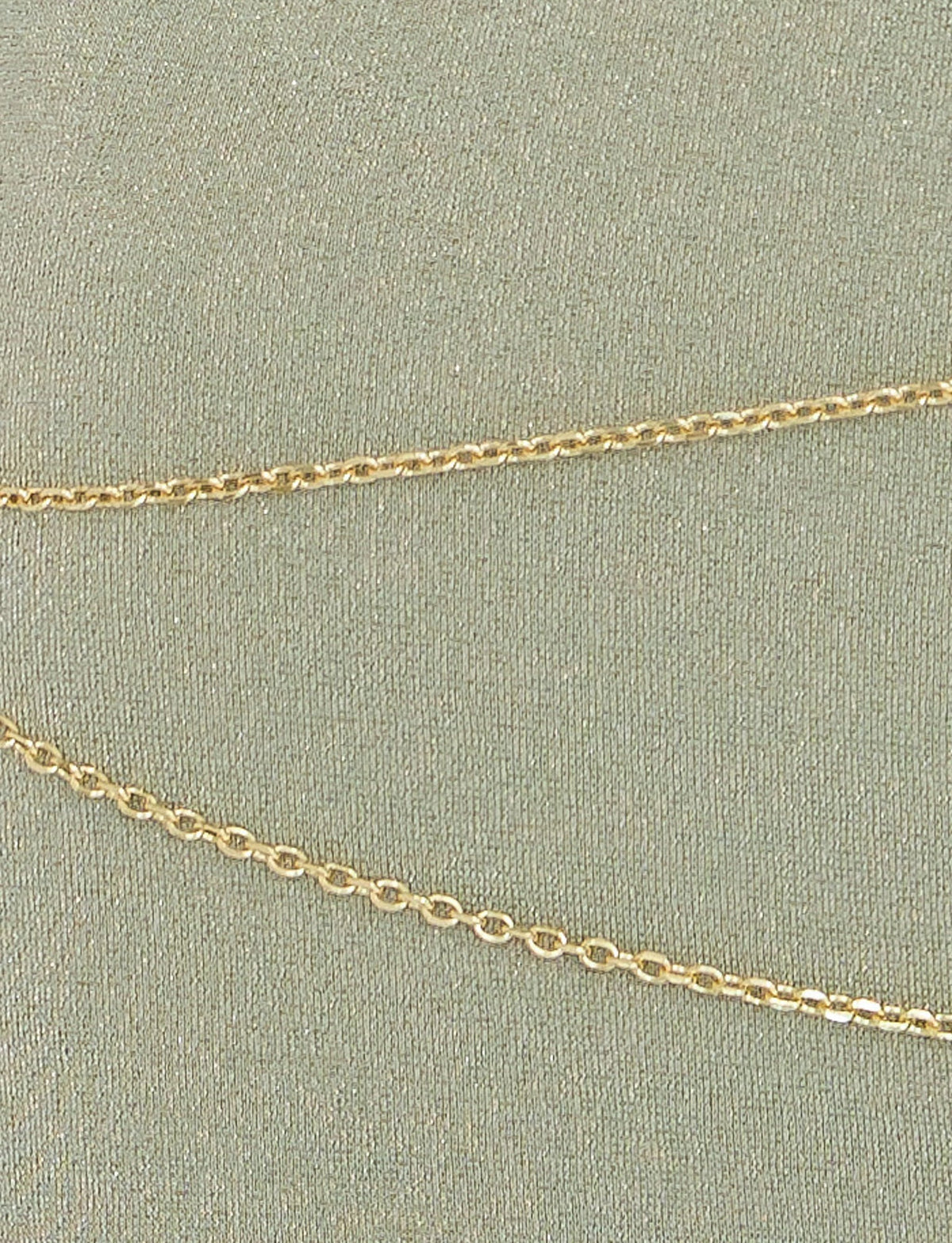 Close-up of a delicate gold chain necklace laid on the textured, light green fabric background of the TIGERMIST OPELI MAXI DRESS in Green Sparkle. The chain features small, linked oval-shaped links that reflect light subtly, adding a touch of elegance reminiscent of an opulent gold belt adorning this exquisite dress.