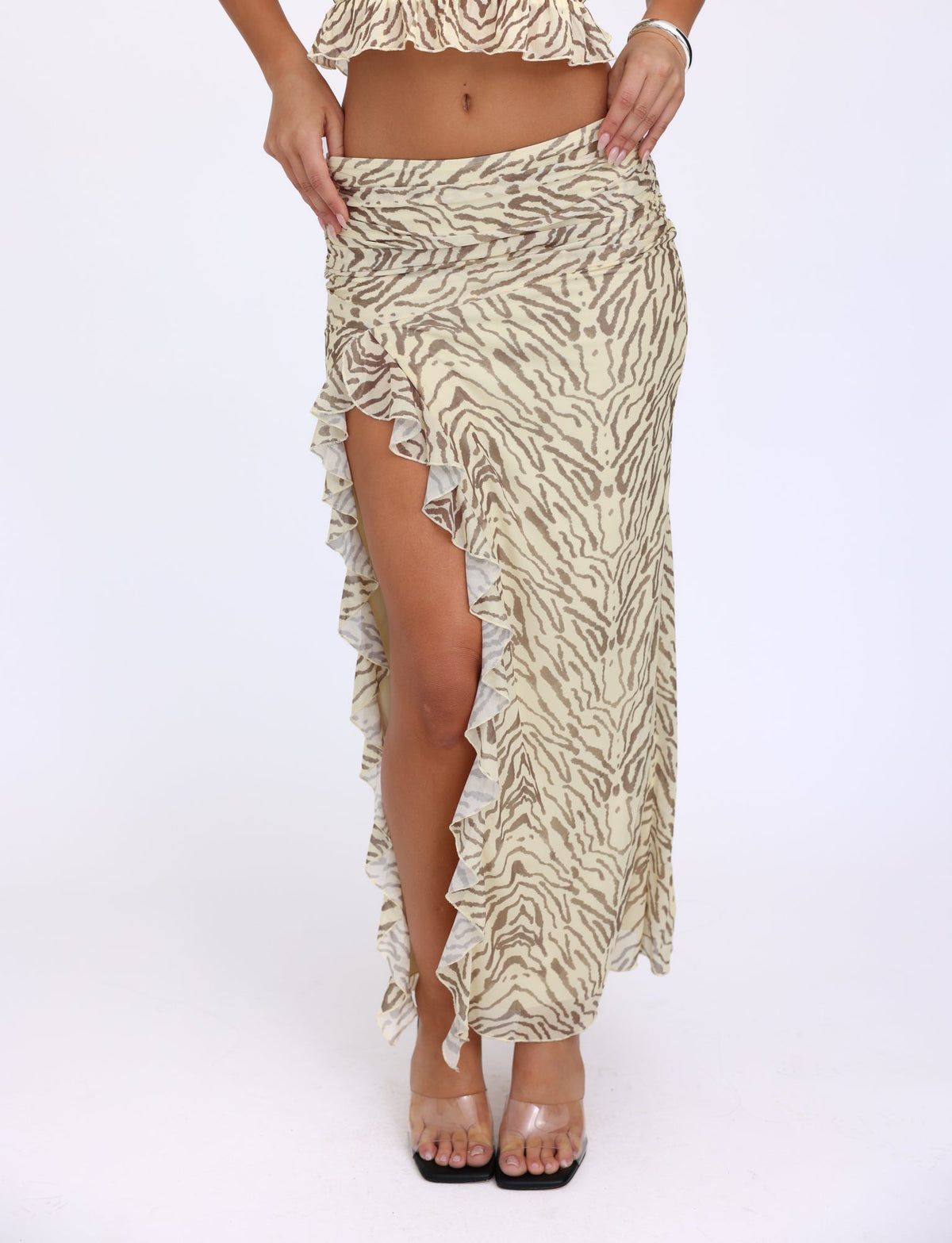 An individual showcasing the RUMER SKIRT - YELLOW : ZEBRA by TIGERMIST, a yellow zebra print maxi skirt with a high split on one side, featuring a subtle ruffle detail along the edge of the slit. They are wearing clear heels, revealing their midriff and part of their legs.