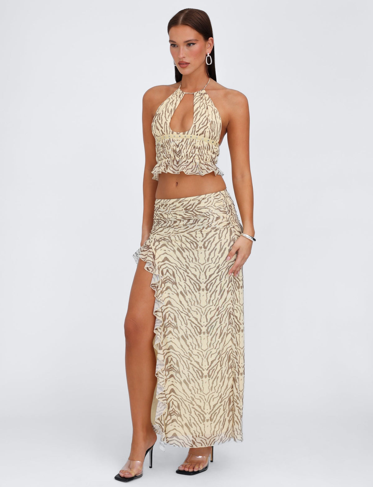 A woman stands posing against a plain background wearing a beige and brown animal print halter top paired with the TIGERMIST RUMER SKIRT in yellow zebra print, which features a high-slit and low-rise waist. She accessorizes with hoop earrings and high-heeled sandals.