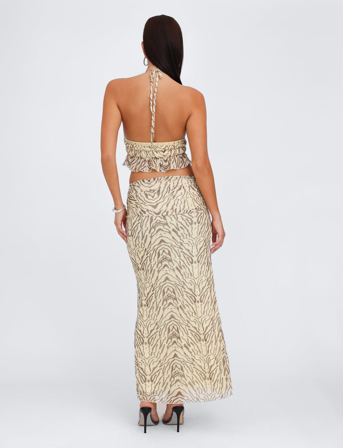 A person with long dark hair stands with their back to the camera, showcasing a backless, halter-neck crop top and the RUMER SKIRT - YELLOW : ZEBRA by TIGERMIST, a maxi skirt featuring a high split. They complete the look with black high heels.