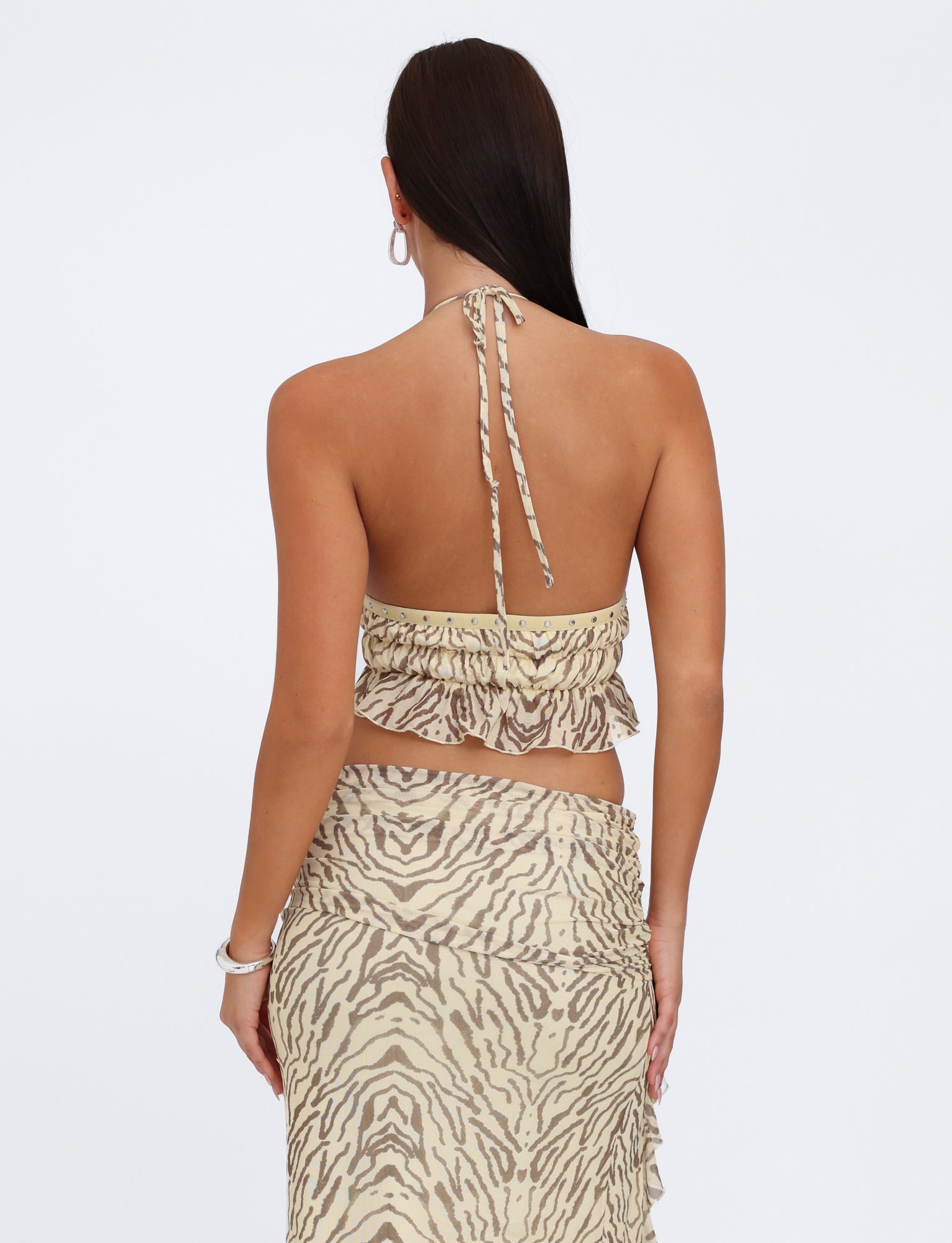 A woman with long dark hair is shown from the back, wearing the RUMER TOP - YELLOW : ZEBRA and a matching skirt from TIGERMIST. The top, featuring adjustable bust panels and ruffled detailing, is tied around her neck, while the skirt wraps around her waist with a ruffled hem. She accessorizes with hoop earrings and a bracelet.