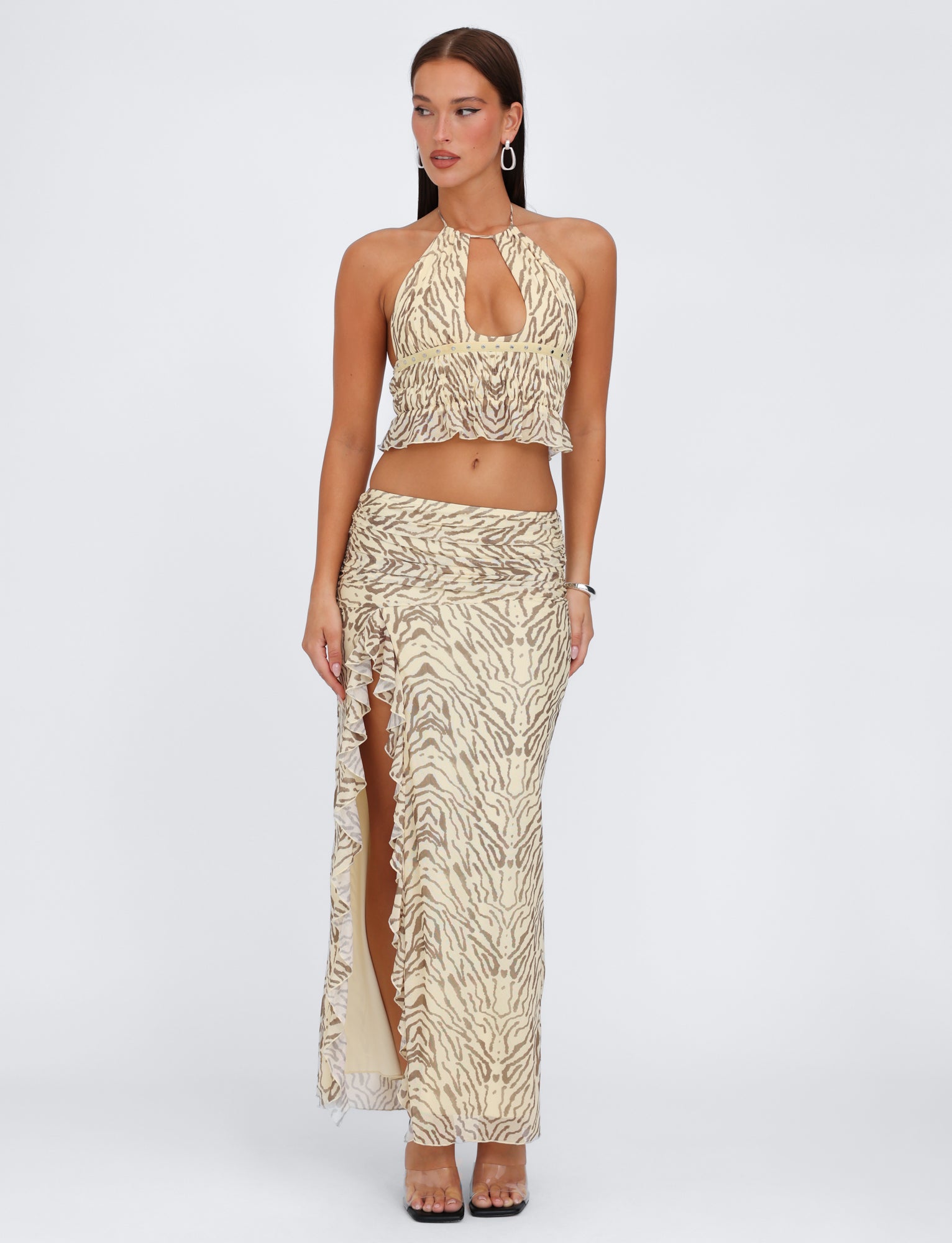 A woman is standing against a plain white background, wearing the RUMER TOP - YELLOW : ZEBRA from TIGERMIST, featuring a beige and brown animal print with adjustable bust panels. She pairs it with a matching high-waisted maxi skirt that boasts a ruffled hem and a high slit. Her look is completed with earrings, bracelets, and high-heeled shoes.