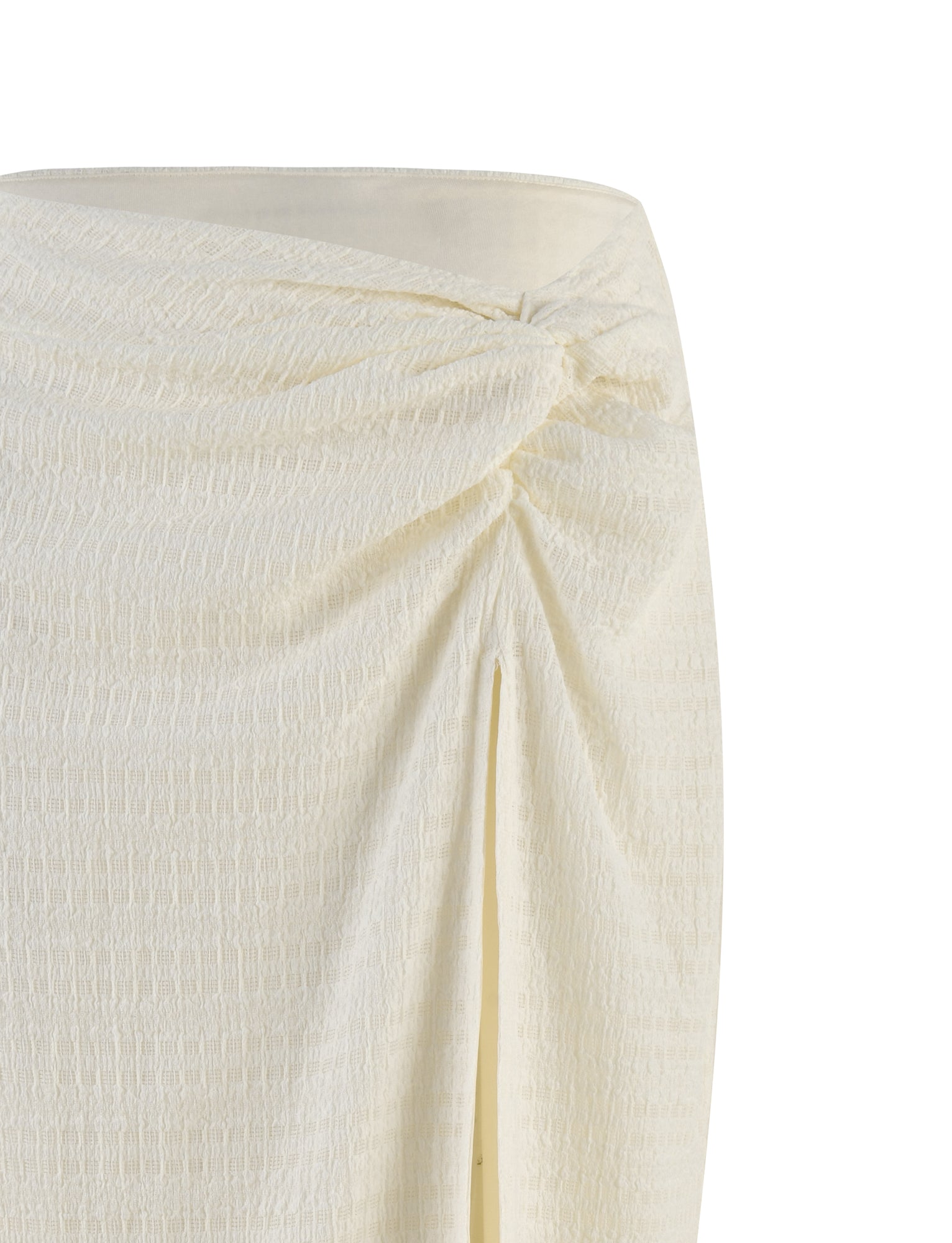 Close-up of the ALDRIDGE SKIRT - WHITE by TIGERMIST. This light-colored textured jersey maxi skirt showcases an asymmetrical twisted knot detail near the waist that creates a gathered effect, along with a front slit revealing a glimpse of fabric underneath. The soft material features a subtle pattern, providing a relaxed silhouette.