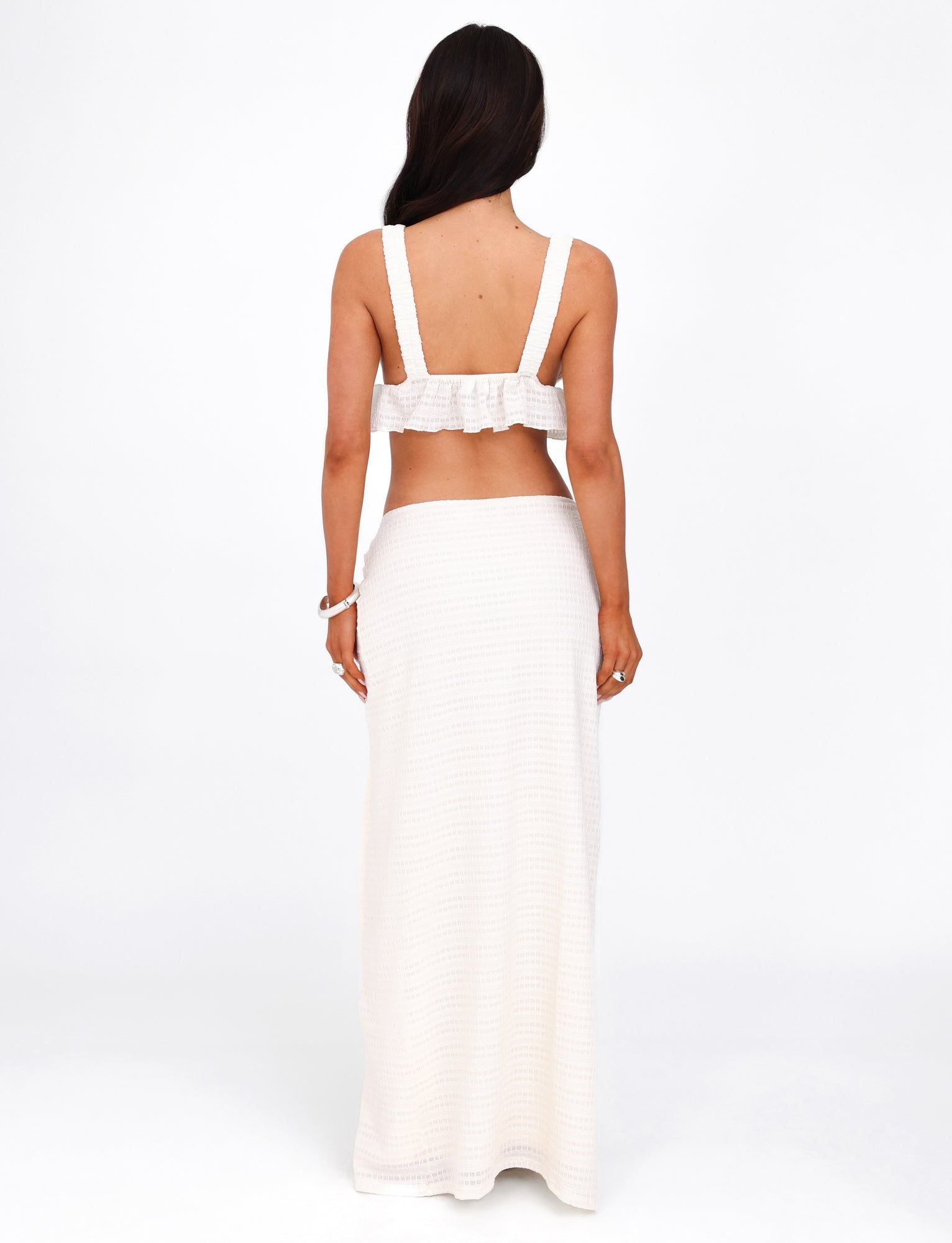 A woman with long dark hair stands facing away, wearing the TIGERMIST ALDRIDGE SKIRT in white, styled with a matching two-piece crochet outfit. The top features wide straps and an exposed back, while the white maxi skirt reaches down to her feet. She accessorizes with a bracelet on her wrist and is photographed against a plain, white background.