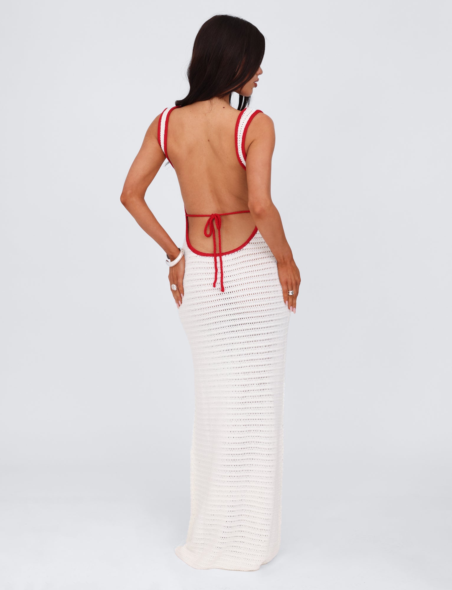 A woman stands with her back to the camera, showcasing the MARLEIGH DRESS - WHITE by TIGERMIST, a stunning long backless white crochet dress with red accents and a halter neck strap. The maxi length dress is set against a plain light grey background.