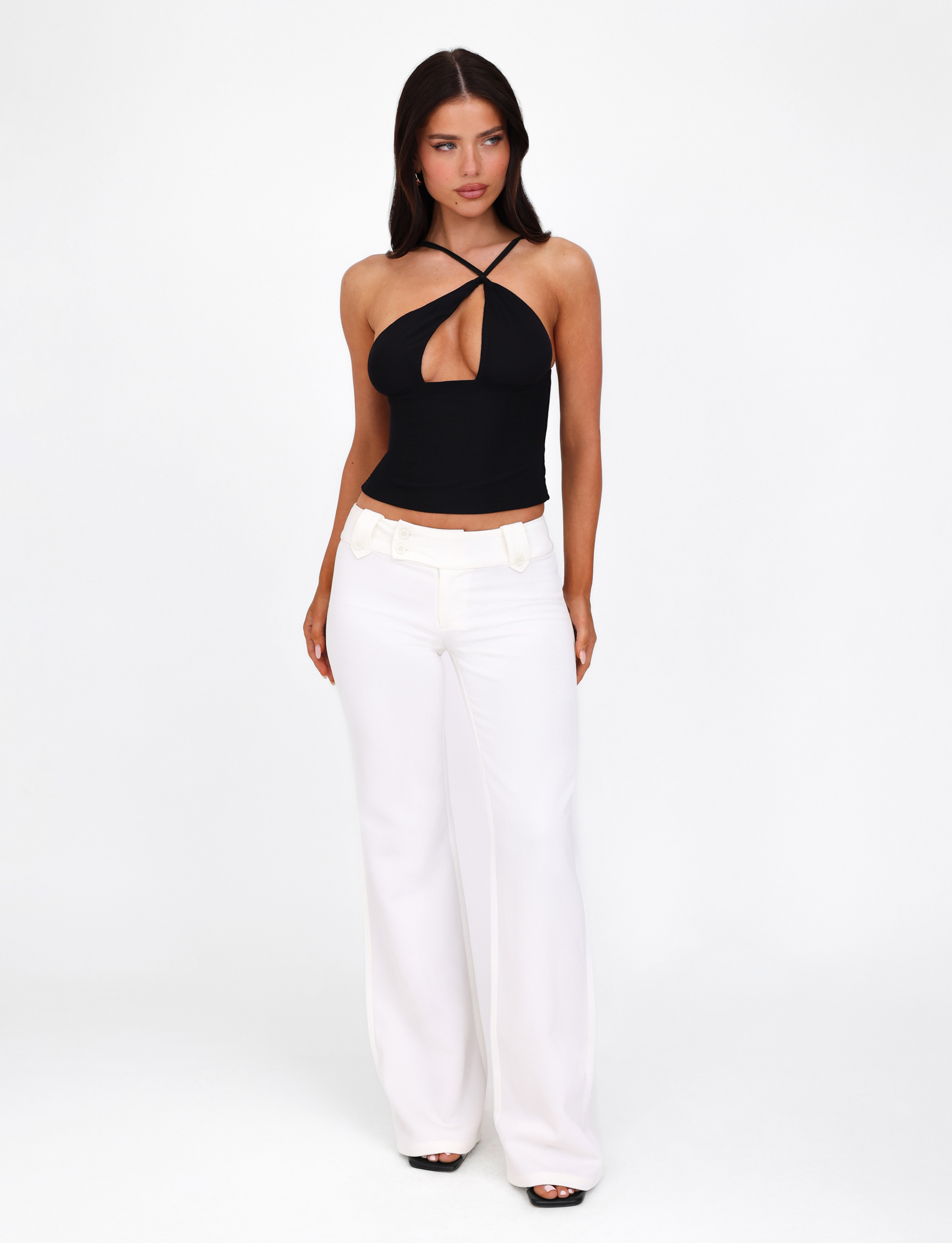 A woman with long dark hair stands against a white background, wearing the TIGERMIST ACADIA TOP in black—featuring mesh fabric and an asymmetric neckline—paired with white wide-leg pants. She looks slightly to the side, hands relaxed at her sides.