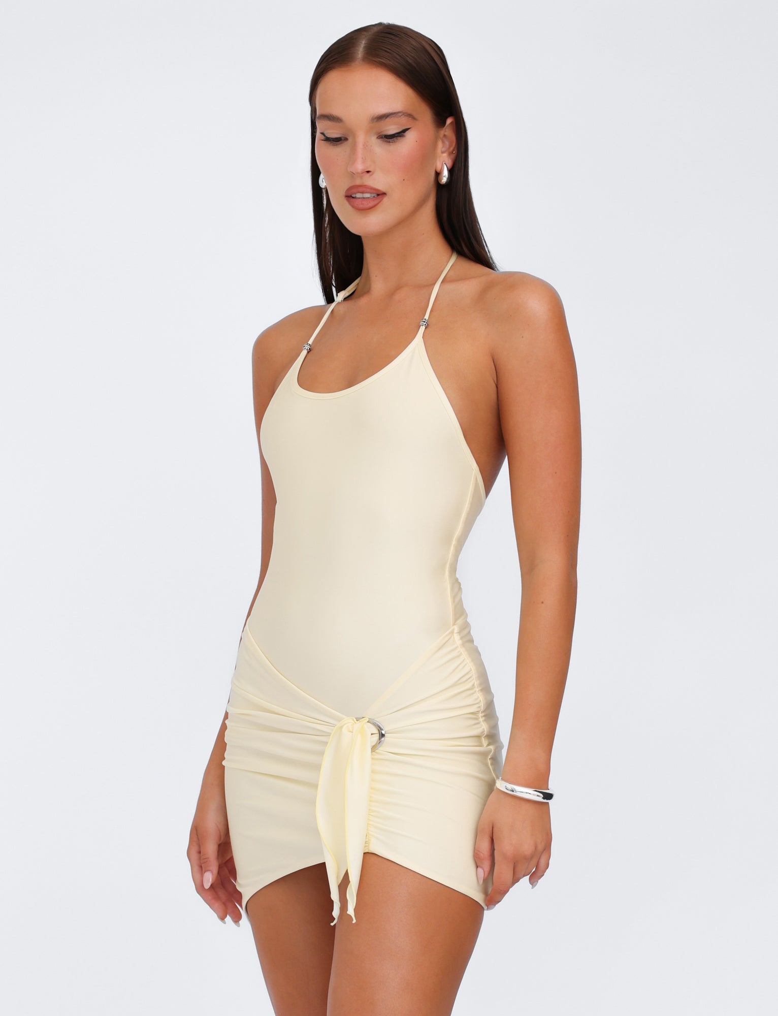 A person with long, straight hair is wearing the ELYSIA DRESS - YELLOW : BUTTER by TIGERMIST, which features a spaghetti strap design, shimmer jersey fabrication, and a tie detail at the waist. They are accessorized with hoop earrings and a bracelet and posed against a plain white background, looking down.