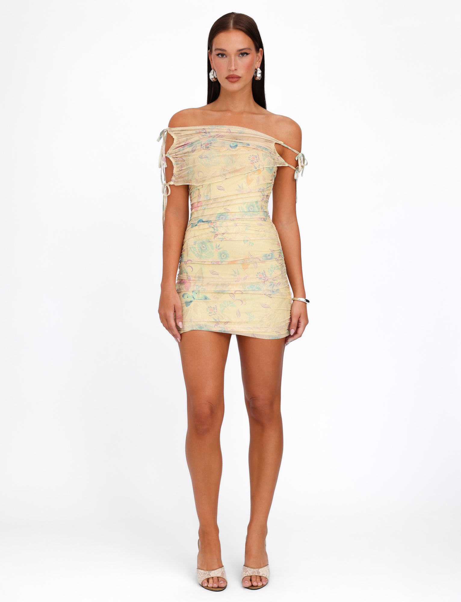 A woman stands against a plain white background, wearing the TIGERMIST KERR DRESS - YELLOW : DELICATE FLORAL, featuring tied straps on the sleeves and ruched detailing. She pairs it with beige high-heeled shoes, minimal jewelry, and has long, straight hair.