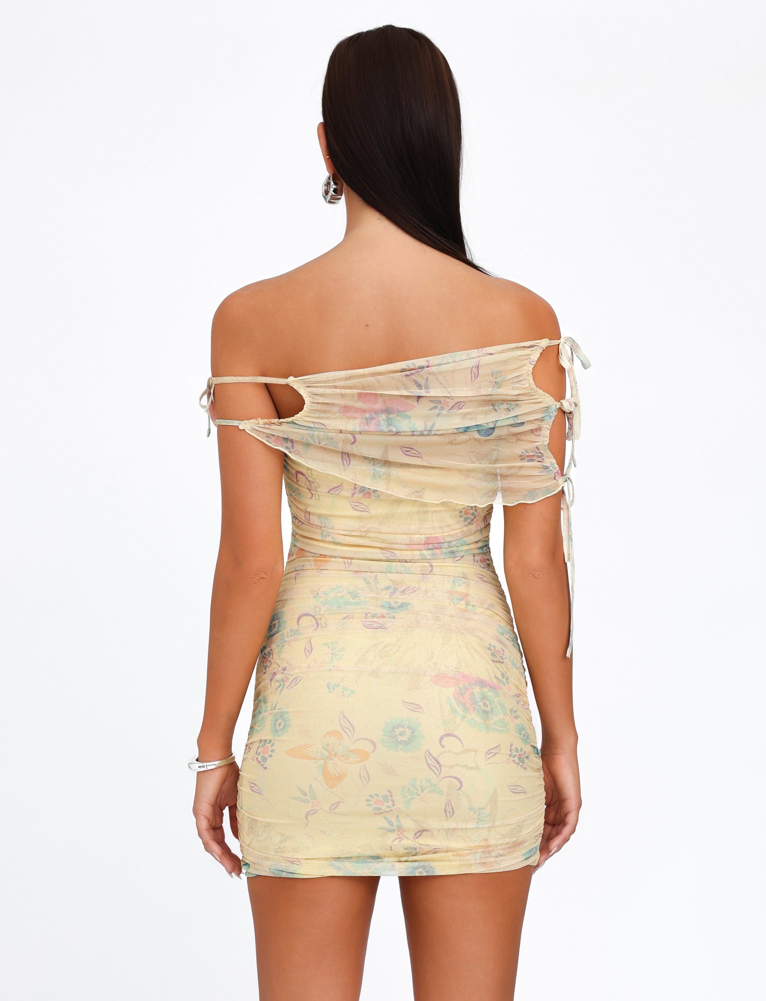 A woman with long brown hair is facing away from the camera, showcasing the KERR DRESS - YELLOW : DELICATE FLORAL by TIGERMIST. The pastel-colored, off-the-shoulder dress features a yellow floral print, lace-up details on the sides, and a fitted silhouette. She accessorizes with hoop earrings and bracelets.