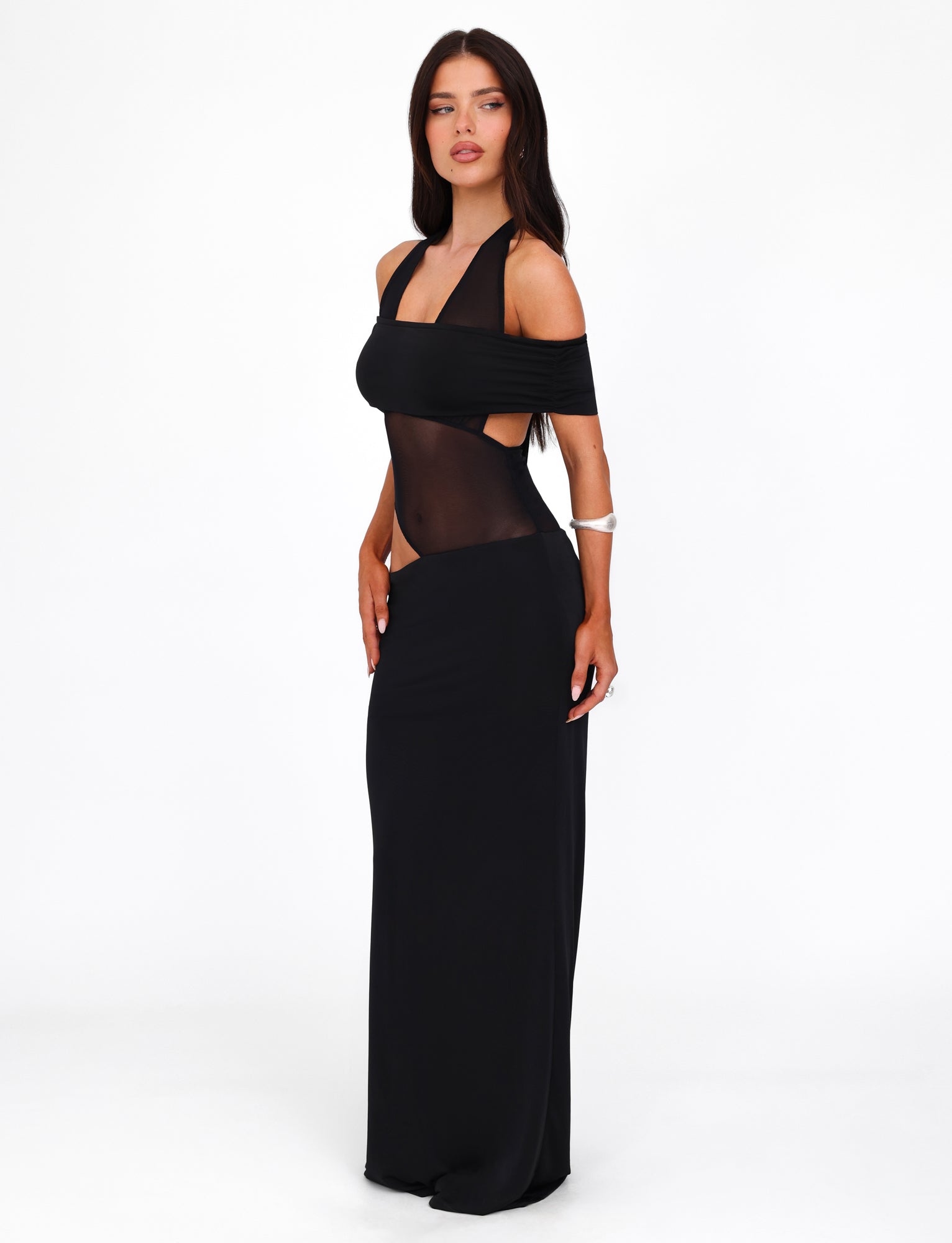 A woman stands against a white background, wearing the EMBERLAIN DRESS - BLACK by TIGERMIST. The maxi-length dress features a one-shoulder neckline, sheer mesh panel midsection, and fitted silhouette. She is posing with one hand on her hip, looking to the side.