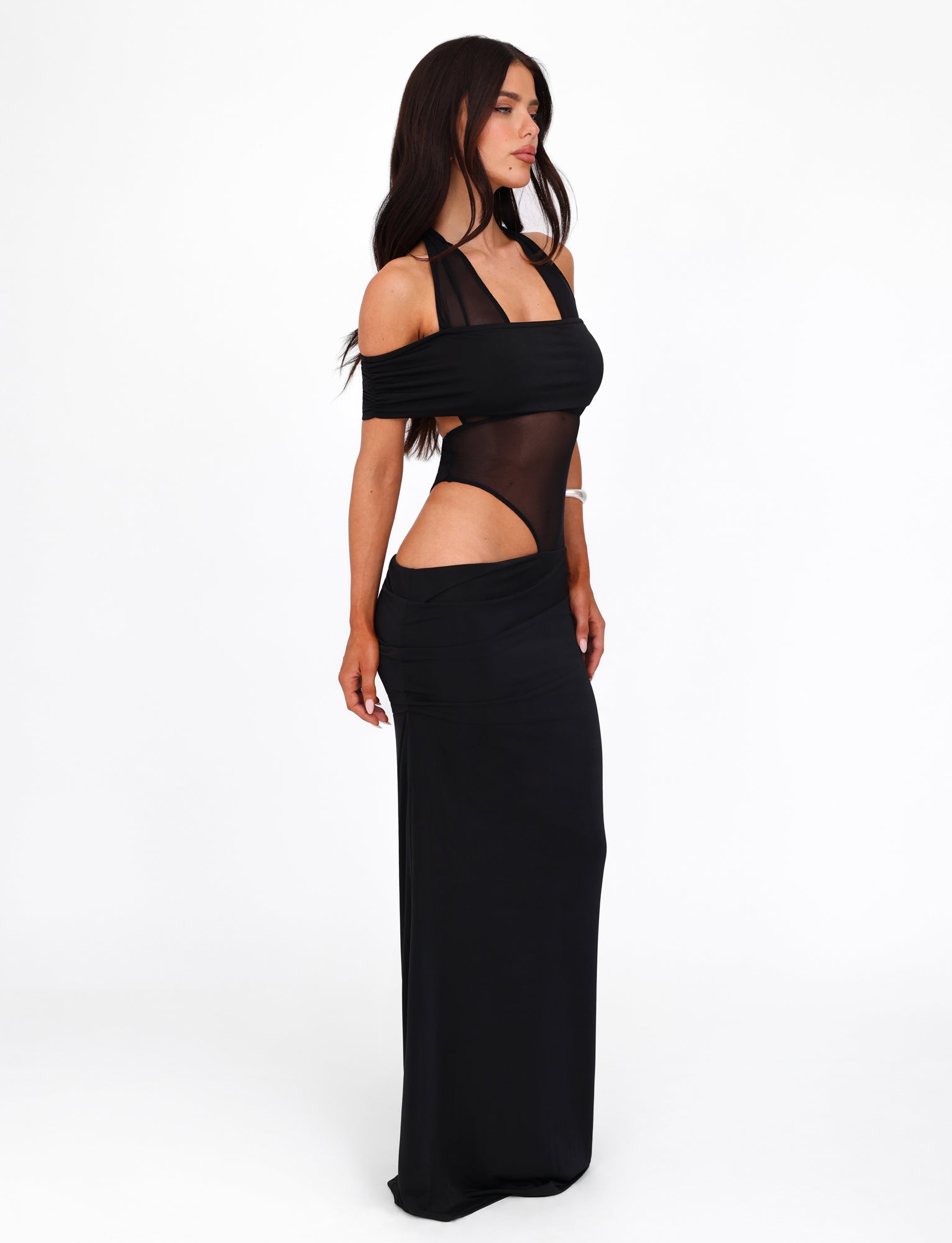 A woman with long dark hair is wearing the elegant TIGERMIST EMBERLAIN DRESS - BLACK, which features a sheer mesh panel bodice with cut-out details on the sides and shoulders, as well as a halter neck design. She stands against a white background, posing sideways with a confident expression.