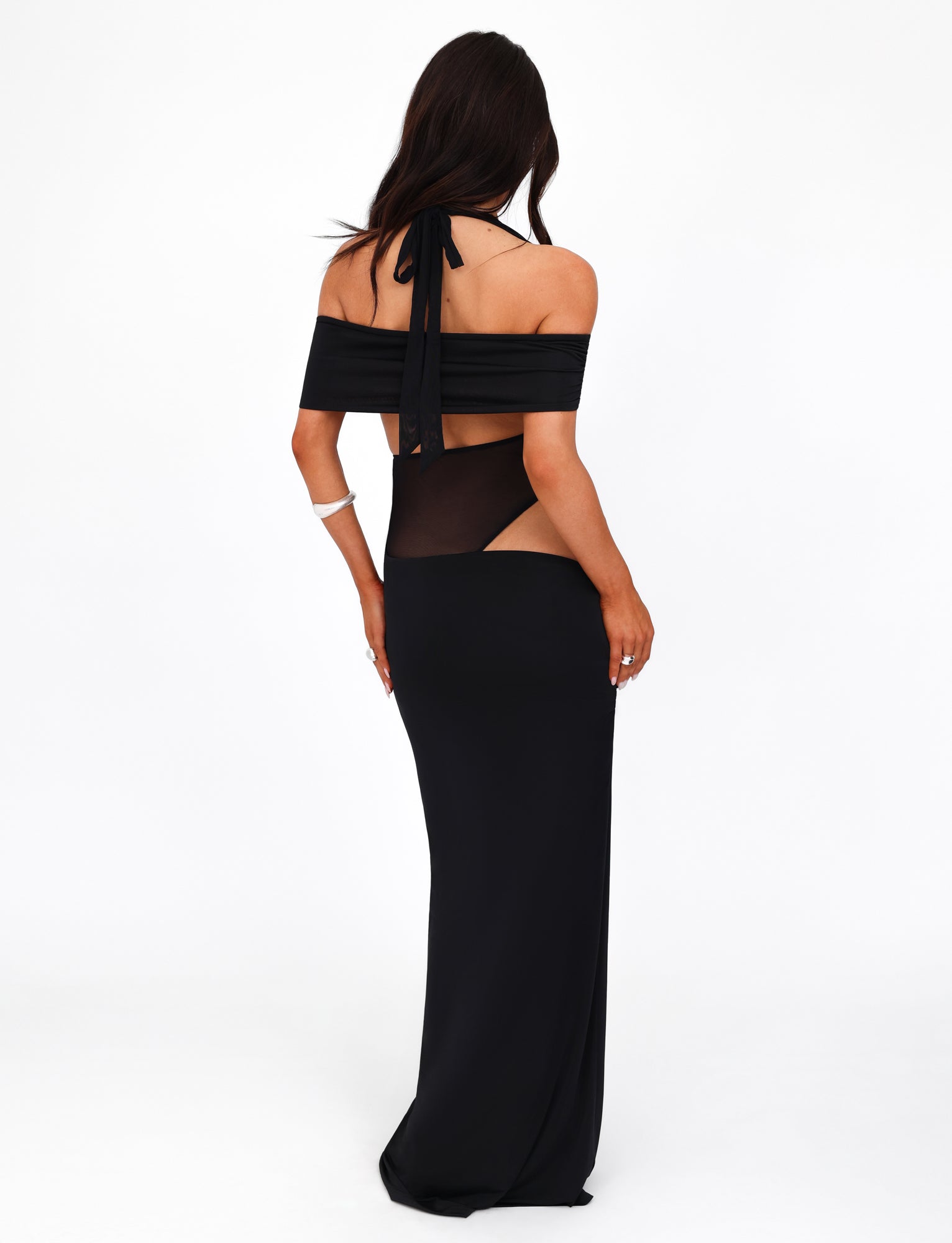 A woman with long dark hair is wearing the TIGERMIST EMBERLAIN DRESS - BLACK, featuring cut-out details at the waist and shoulder straps. She is facing away, showcasing the back of the maxi-length dress against a plain white backdrop.
