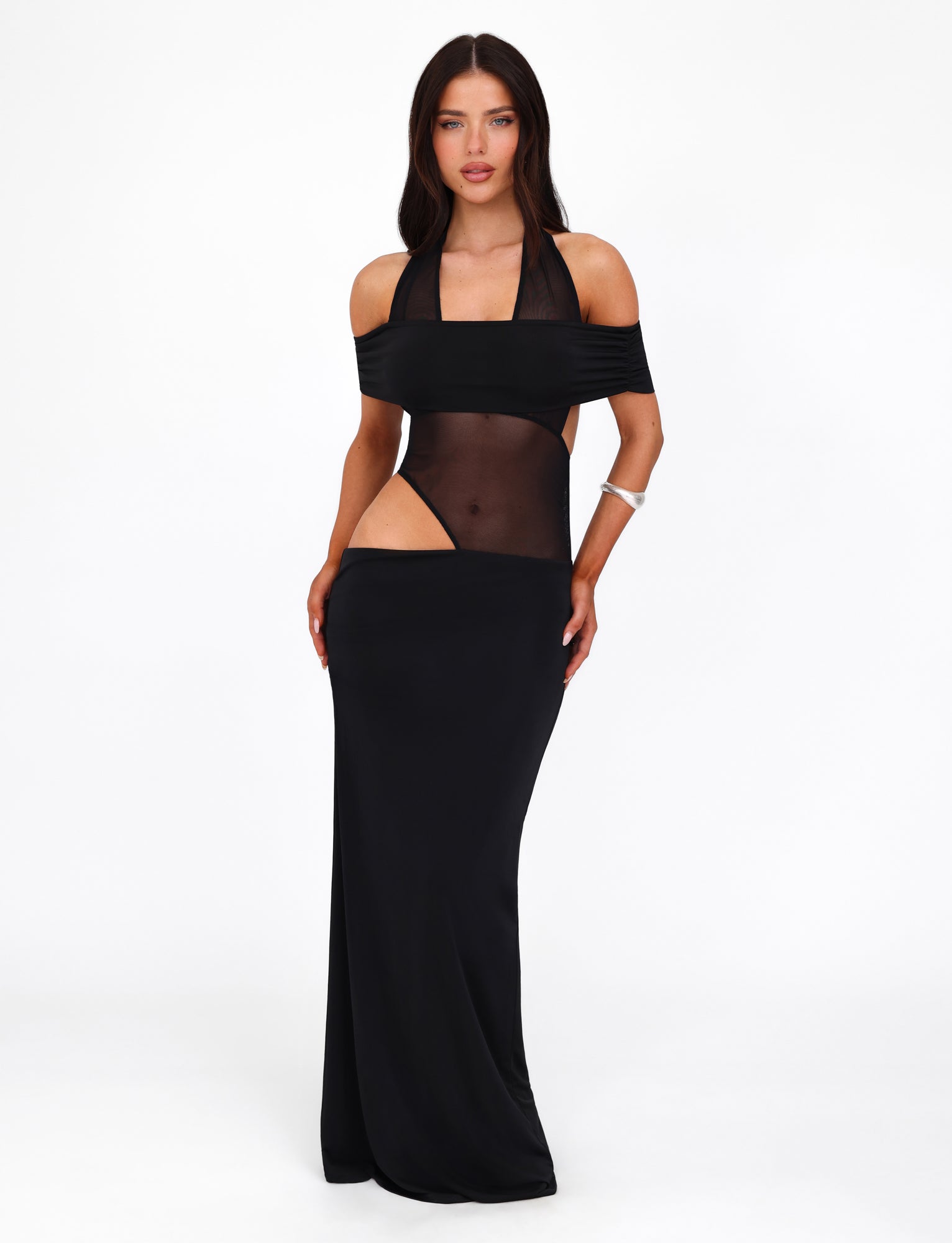 Against a white backdrop stands a woman donning the EMBERLAIN DRESS by TIGERMIST. The black dress features a halter neckline and a mesh panel that offers an alluring glimpse of skin. Her long, dark hair flows past her shoulders, perfectly complementing the dress's maxi length. She accessorizes with a silver bracelet on her right wrist and maintains a neutral expression as her hands rest on her thighs.