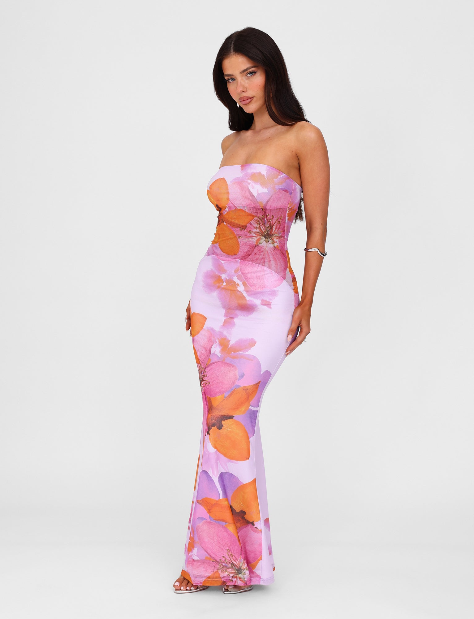 A woman with long dark hair is standing and wearing the Morgan Dress - Pink by Poppy Summer from TIGERMIST, a strapless, form-fitting piece adorned with large, vibrant orange and pink floral patterns. She is looking directly at the camera, with one arm bent and placed on her hip against a plain white background.