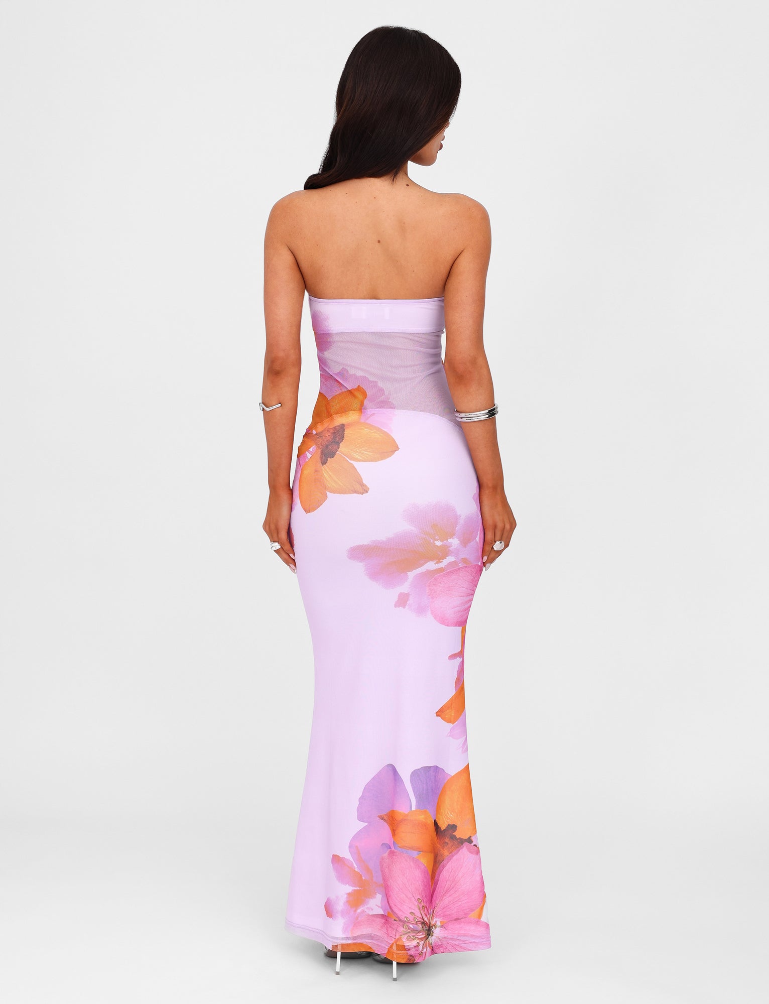 A woman with long brown hair is facing away, showcasing the MORGAN DRESS - PINK from the TIGERMIST By Poppy Summer collection. The strapless, floor-length lilac gown features large, vibrant orange and pink floral print designs. She completes her look with a silver bracelet against a plain white background.