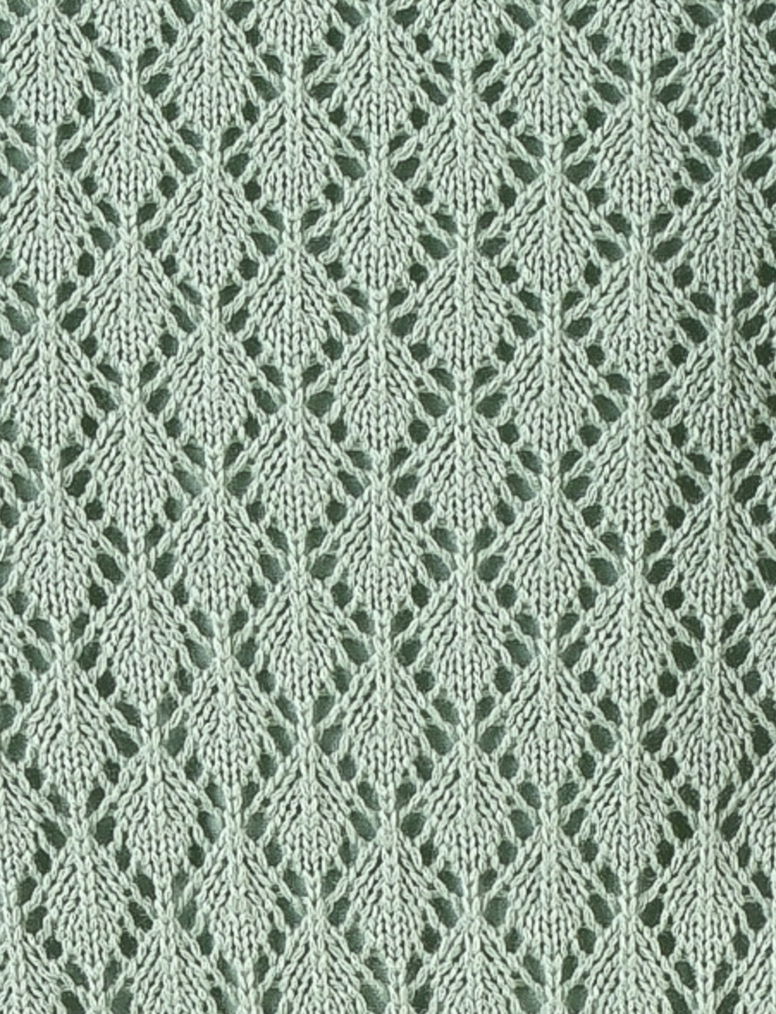 A close-up of the COLLINA SKIRT - GREEN from TIGERMIST reveals a light green knitted fabric showcasing a repeating leaf pattern with intricate lacework, reminiscent of the delicate details found in crochet skirts. The design is uniform, creating a textured and elegant appearance. The background features a slightly darker green hue, which enhances the contrast and highlights the exquisite pattern.