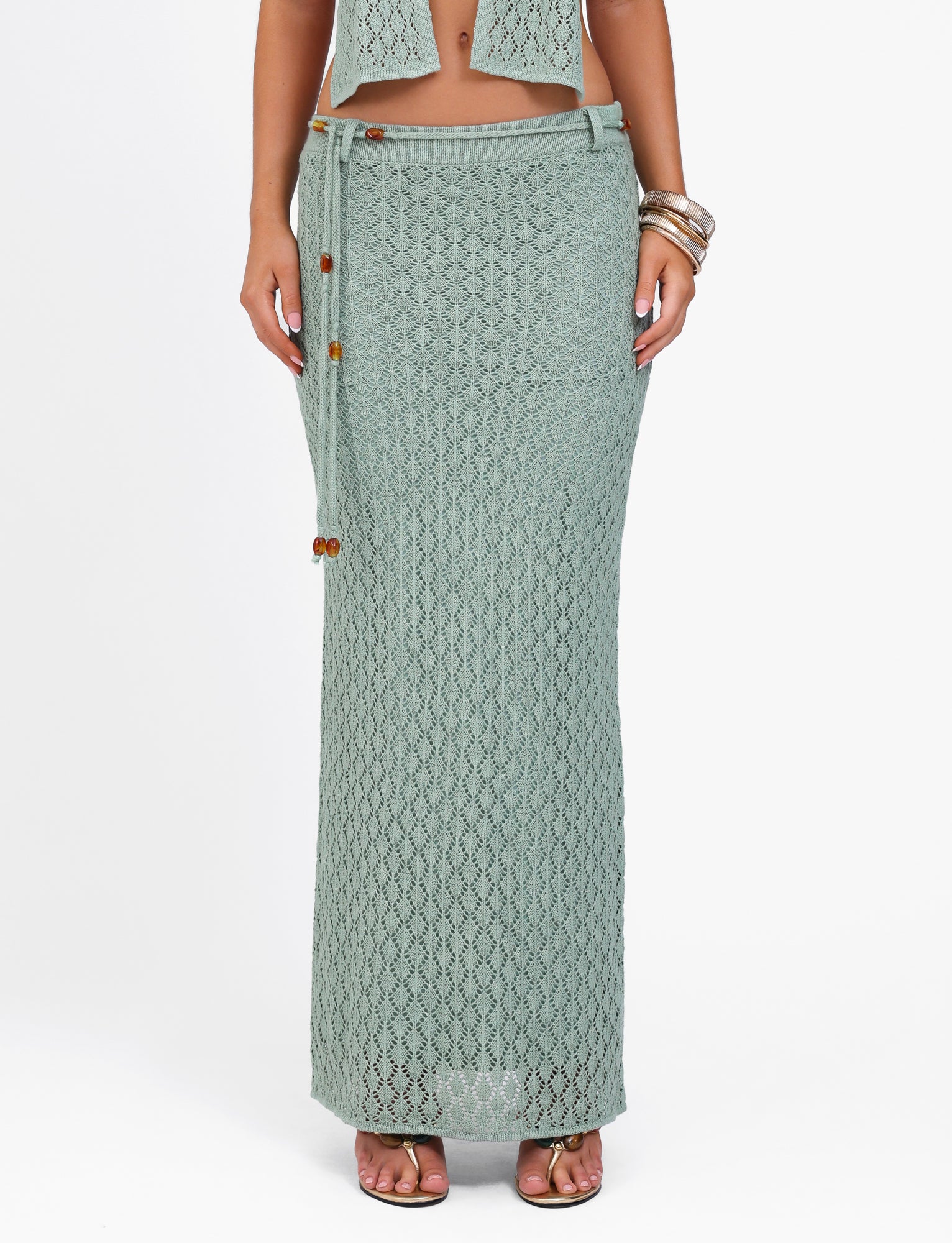 A person standing on a white background wears the stylish COLLINA SKIRT - GREEN by TIGERMIST. The light green crochet maxi skirt with a tie closure and wooden bead details touches the floor. The individual accessorizes with bangles and brown sandals, while their upper body is partially visible.