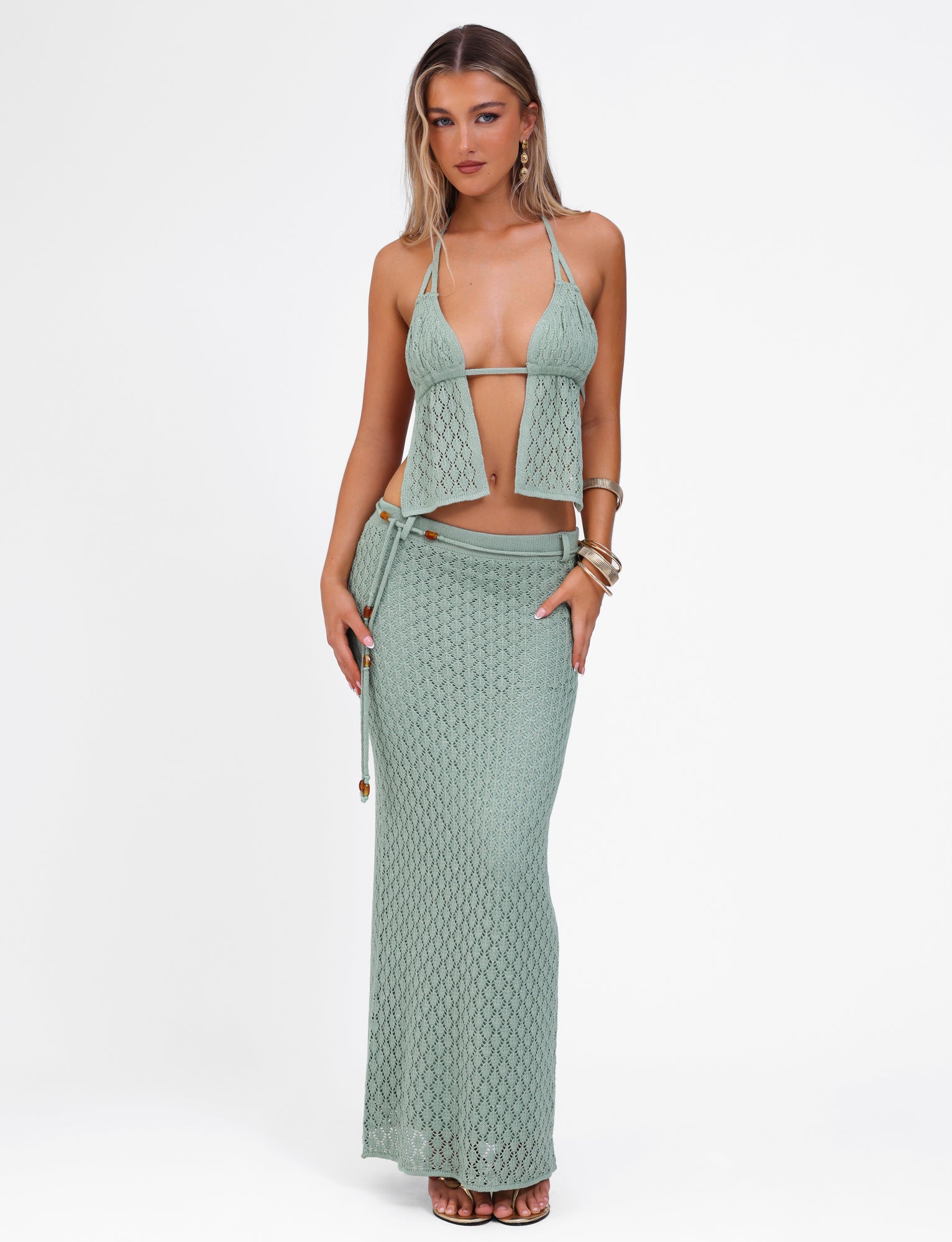 A woman stands against a plain white background wearing the COLLINA SKIRT - GREEN by TIGERMIST, styled with a matching knitted halter top. She accessorizes with bracelets and has long hair styled in loose waves.
